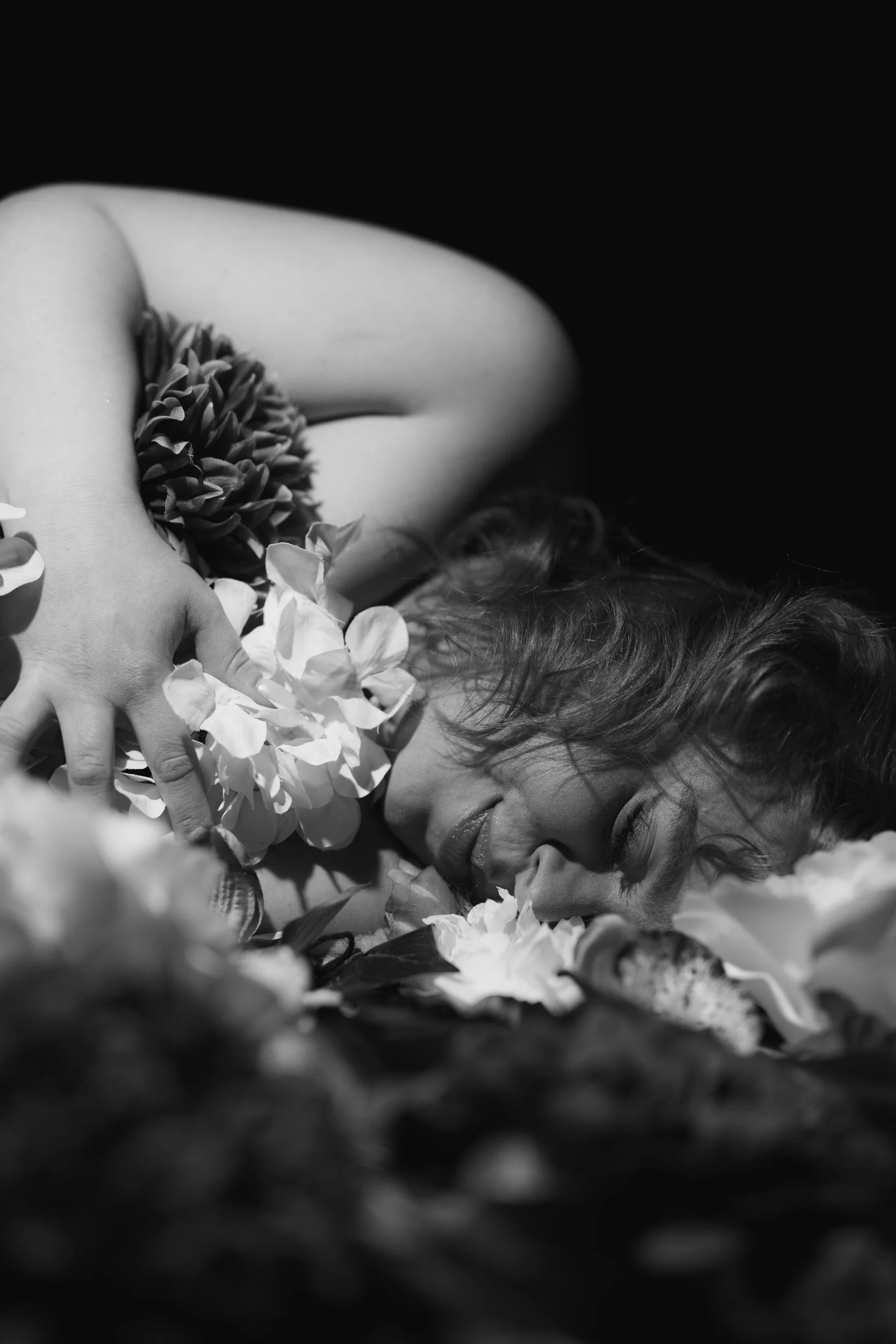 A woman is laying on top of flowers. The image is black and white. She is holding flowers againts her chest and her eyes are closed.