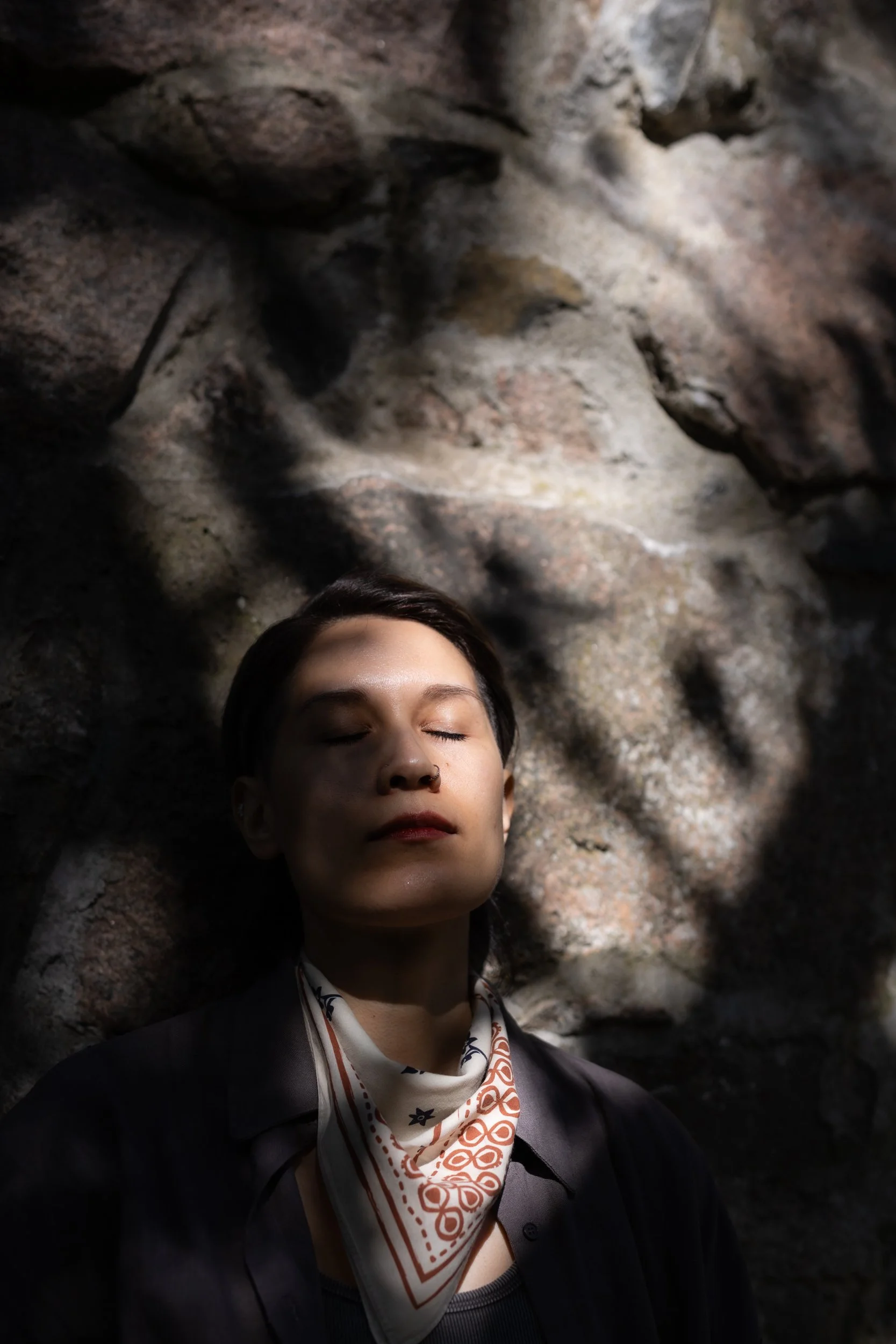 A person with short dark hair and a nose ring, eyes closed, standing against a rocky background, wearing a dark jacket and a patterned bandana around her neck.