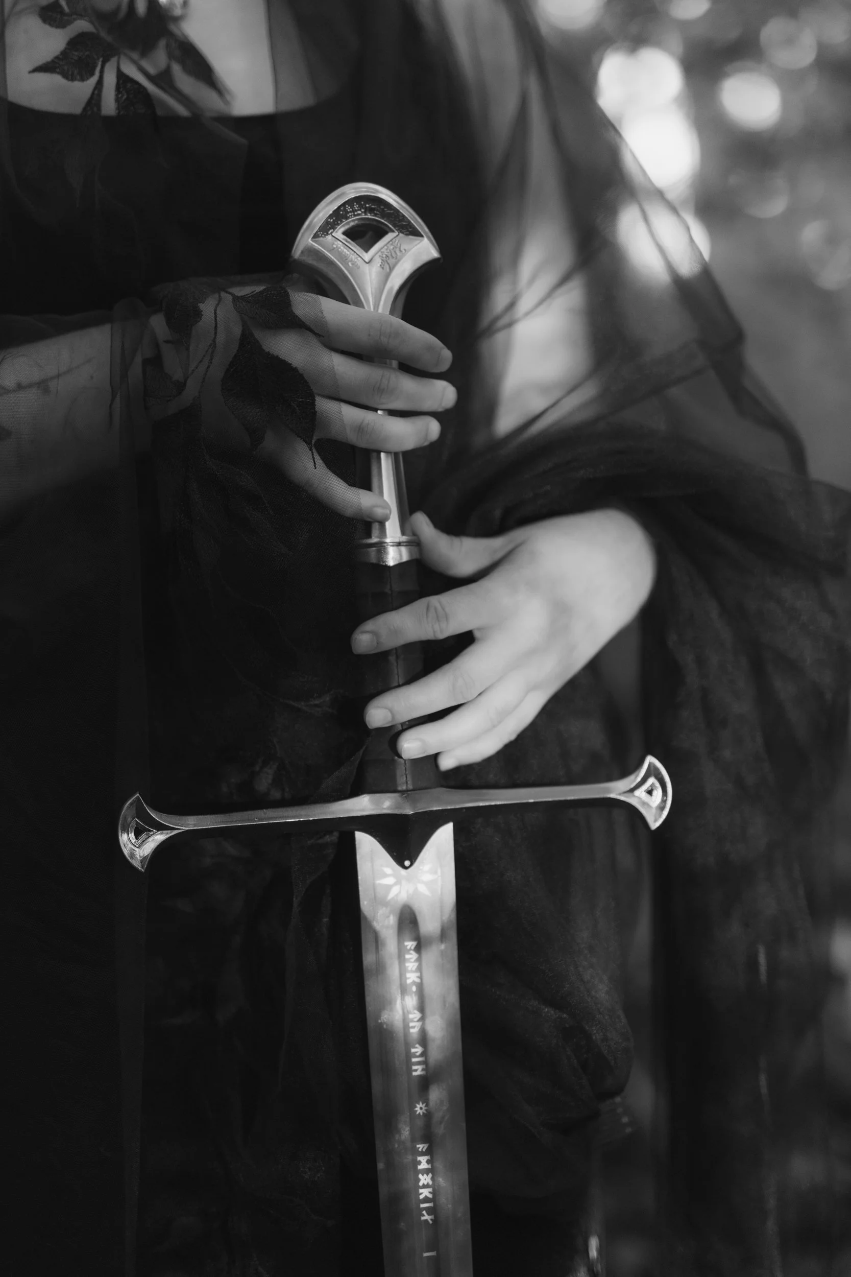 Close-up black-and-white photo of a person's hand holding a medieval style sword with a detailed hilt, the person is wearing sheer black lace gloves and a black top, background blurred with bokeh lights.
