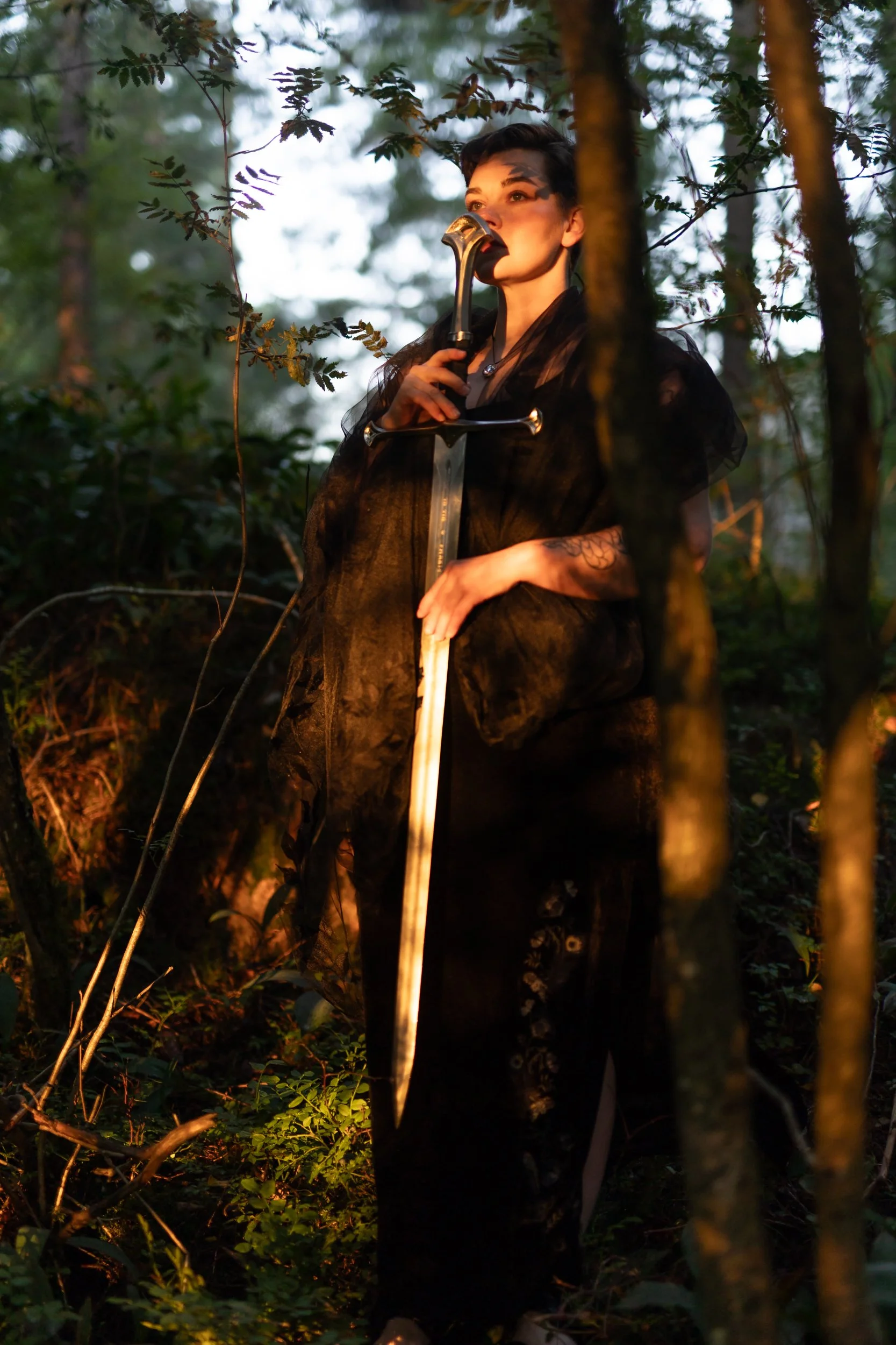 Person with short dark hair, dressed in black, holding a sword in a dense forest during dusk or dawn, surrounded by trees and foliage.