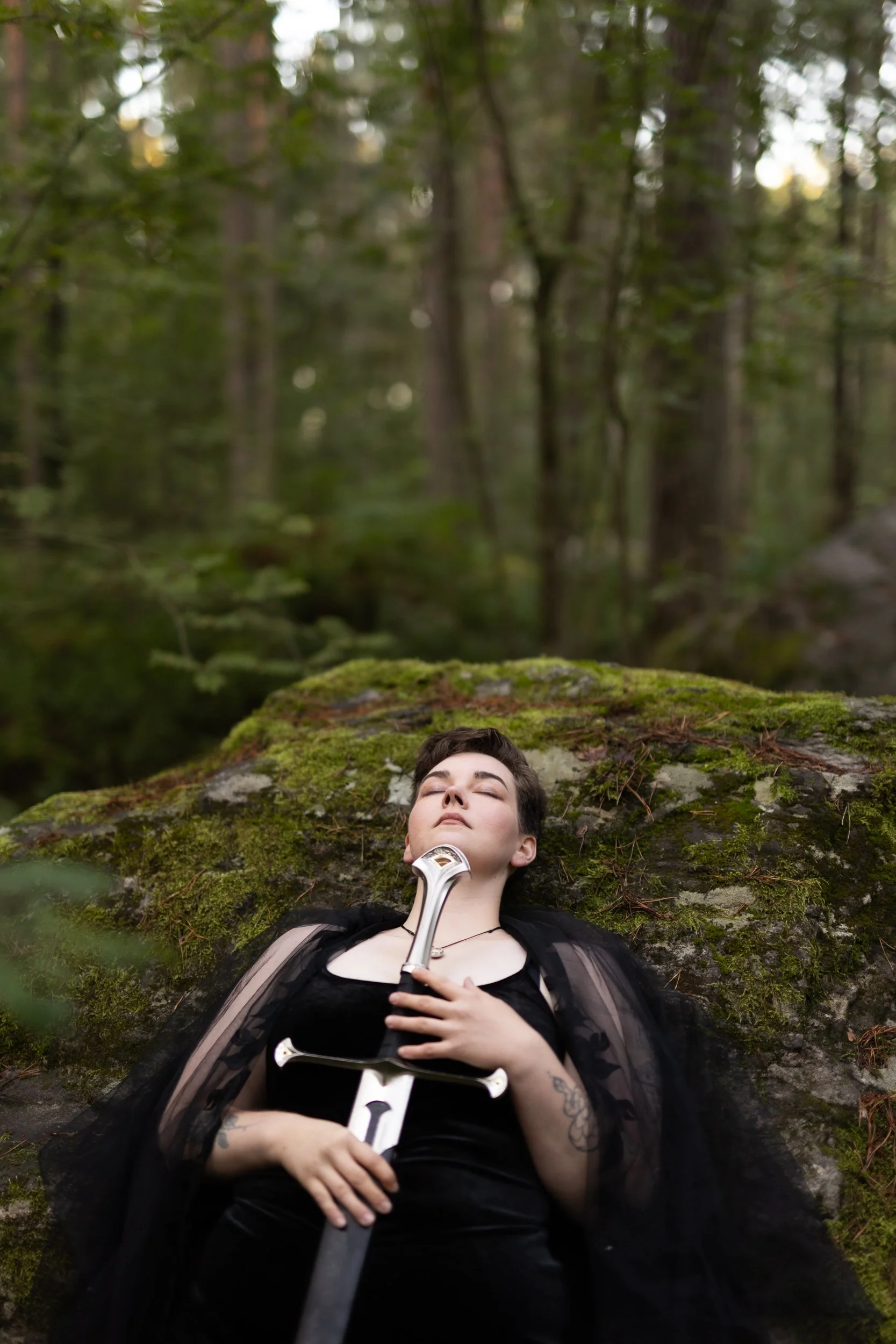 Young person with short dark hair lying on moss-covered ground in a forest, eyes closed, holding a sword across their chest, wearing a black top and sheer outer garment, surrounded by lush green trees.
