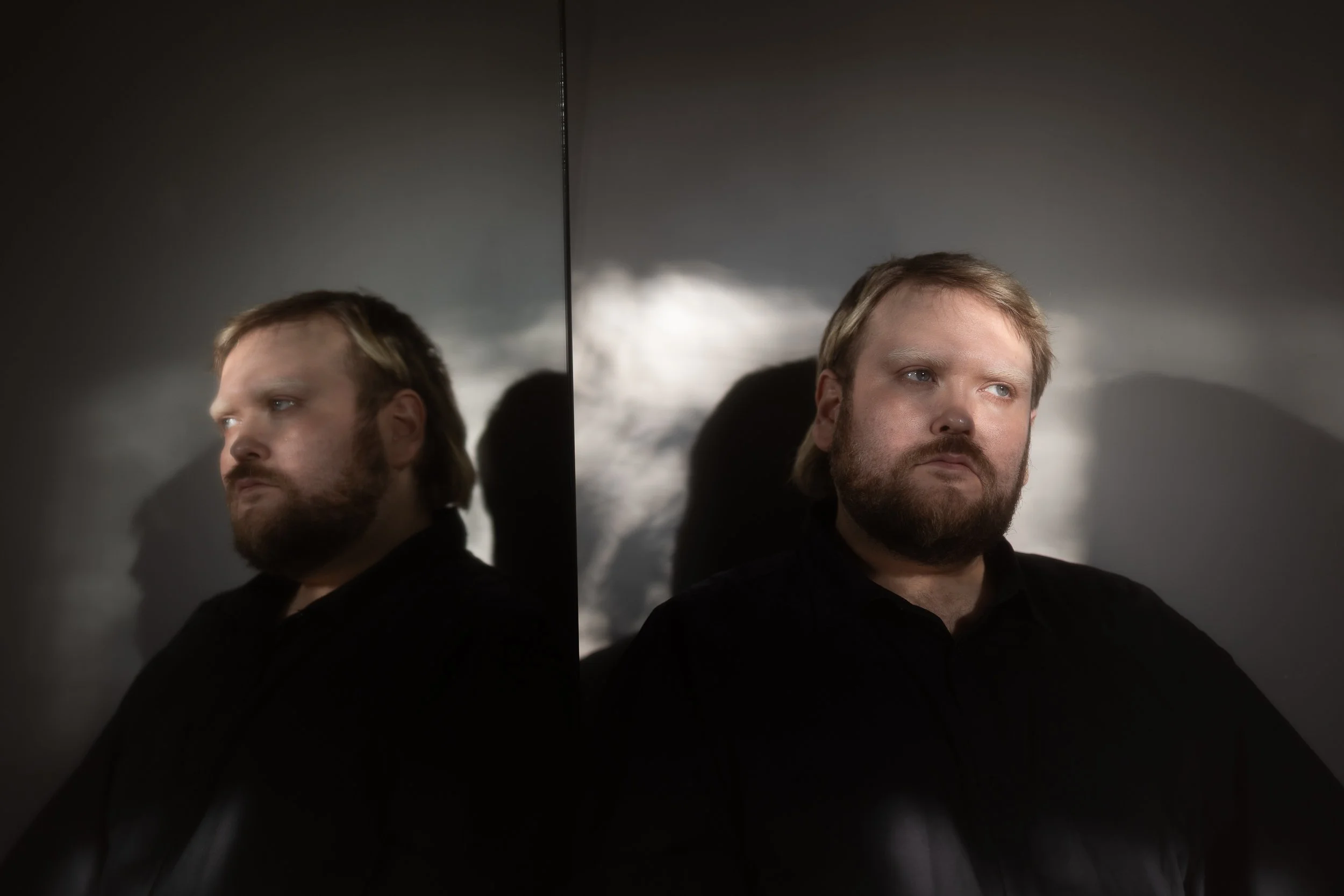 Taneli is leaning on a wall and on their rights side is a mirror. The sun light paints beautiful highlights on Taneli's face. They are looking slightly right of the camera. The mirror reflection duplicates Taneli.