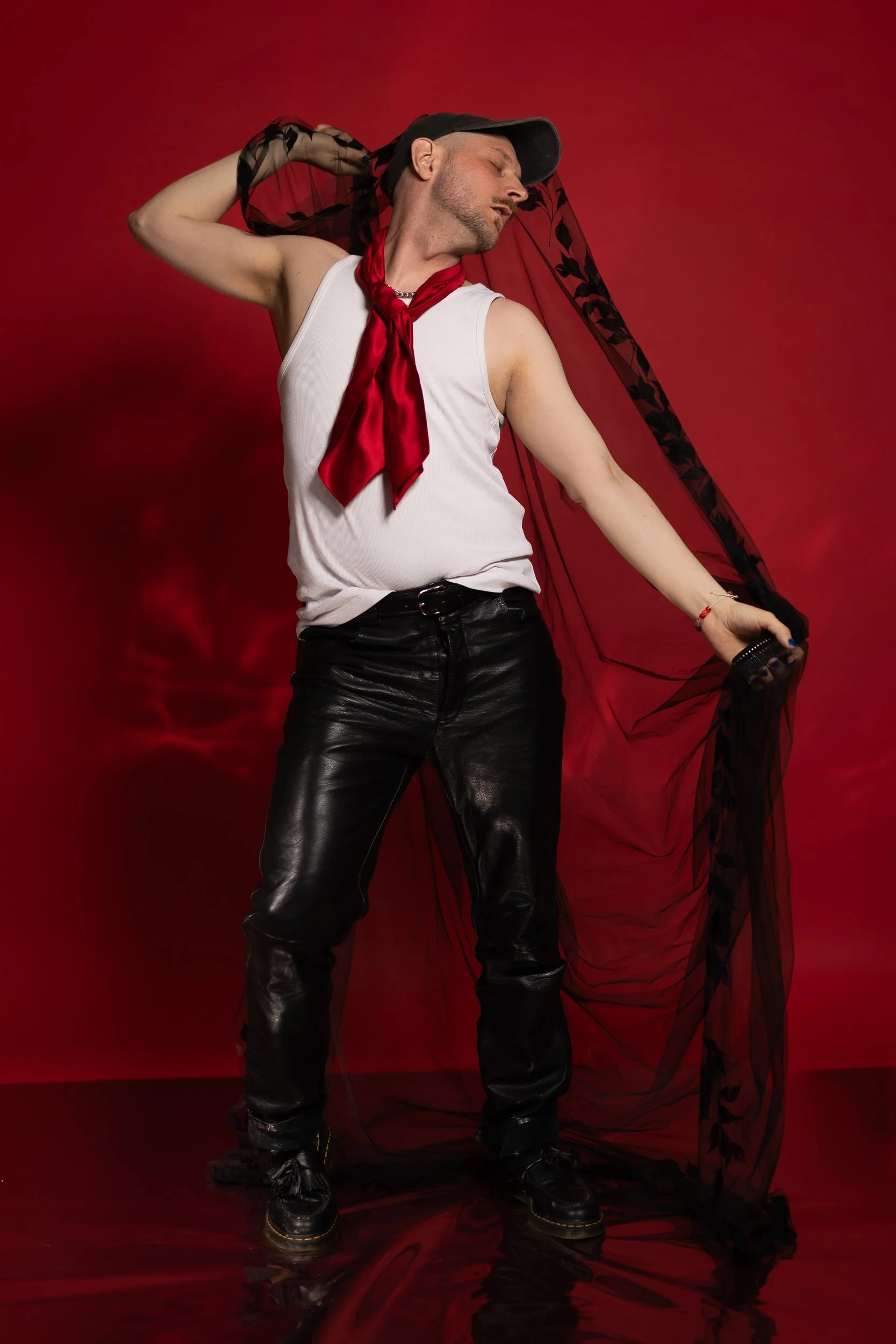 Dramatic and powerfull portrait photo. A person wearing leather pants and white shirt stands in contra posto. They are holding a black veil behind their back.