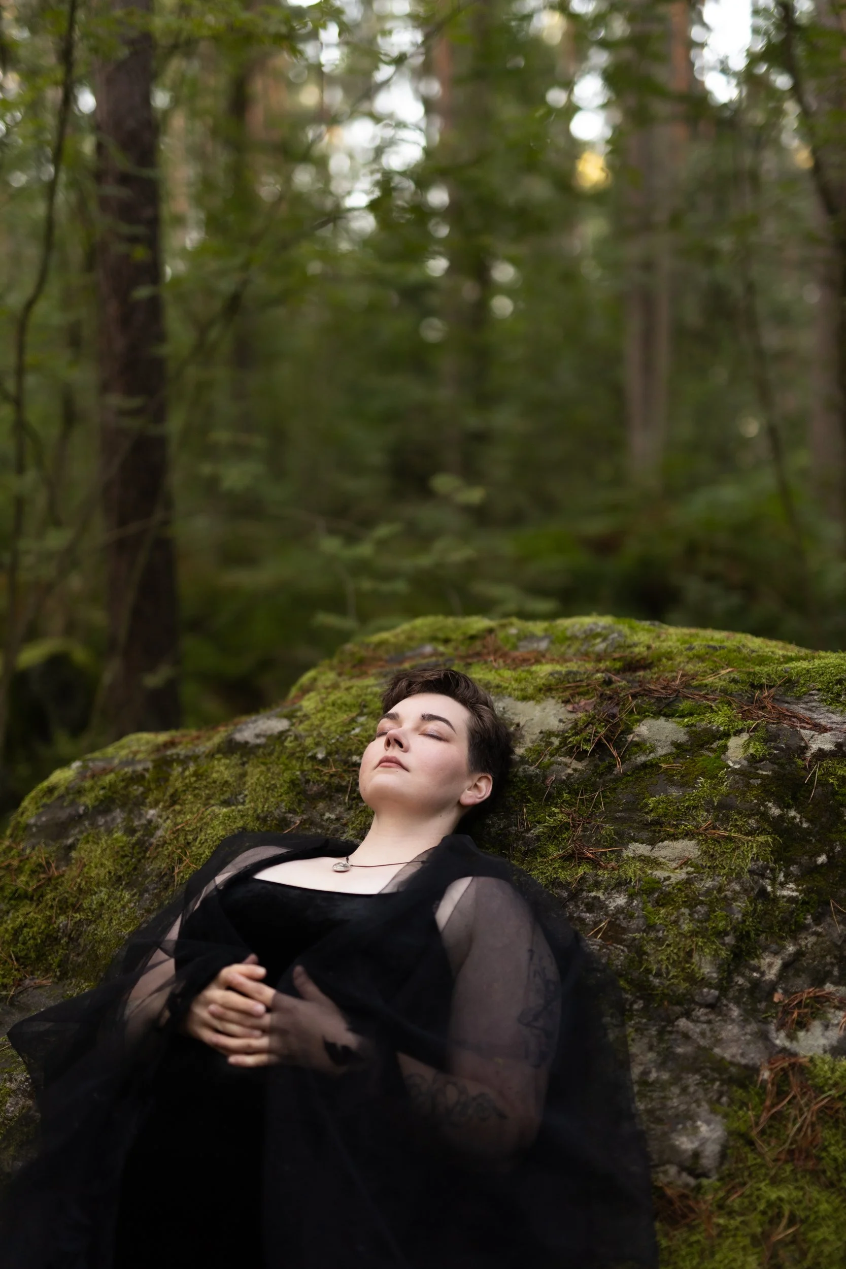 A person with short dark hair and fair skin lying down on mossy forest ground, eyes closed, wearing a black sheer dress and a necklace, surrounded by trees and green foliage.