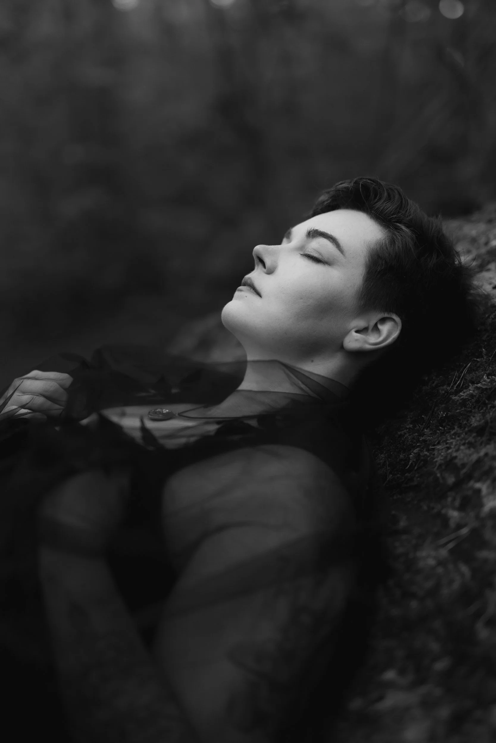 A black and white photo of a person lying down with her eyes closed, appearing peaceful with their head resting on a surface outdoors.