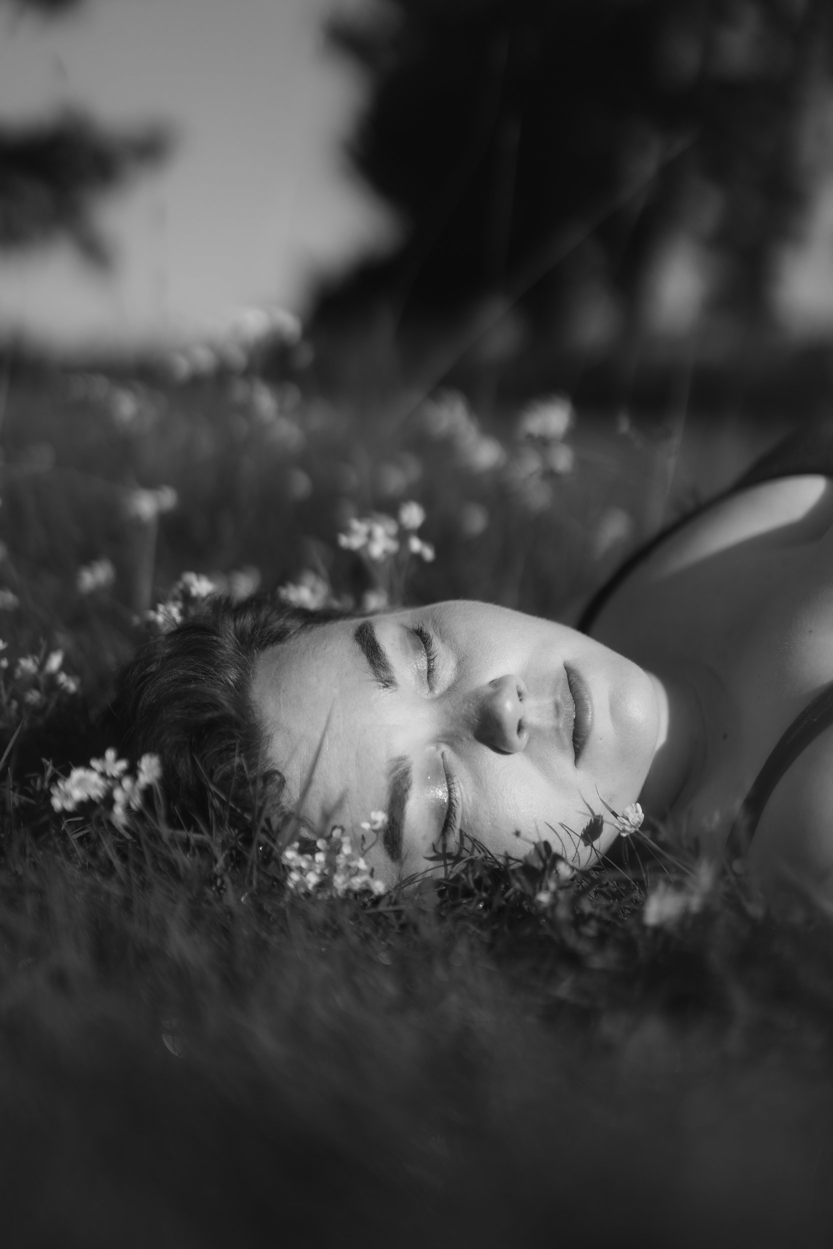 Black and white photo of a person lying in a field of grass and wildflowers with their eyes closed, a peaceful expression, and surrounded by natural elements.