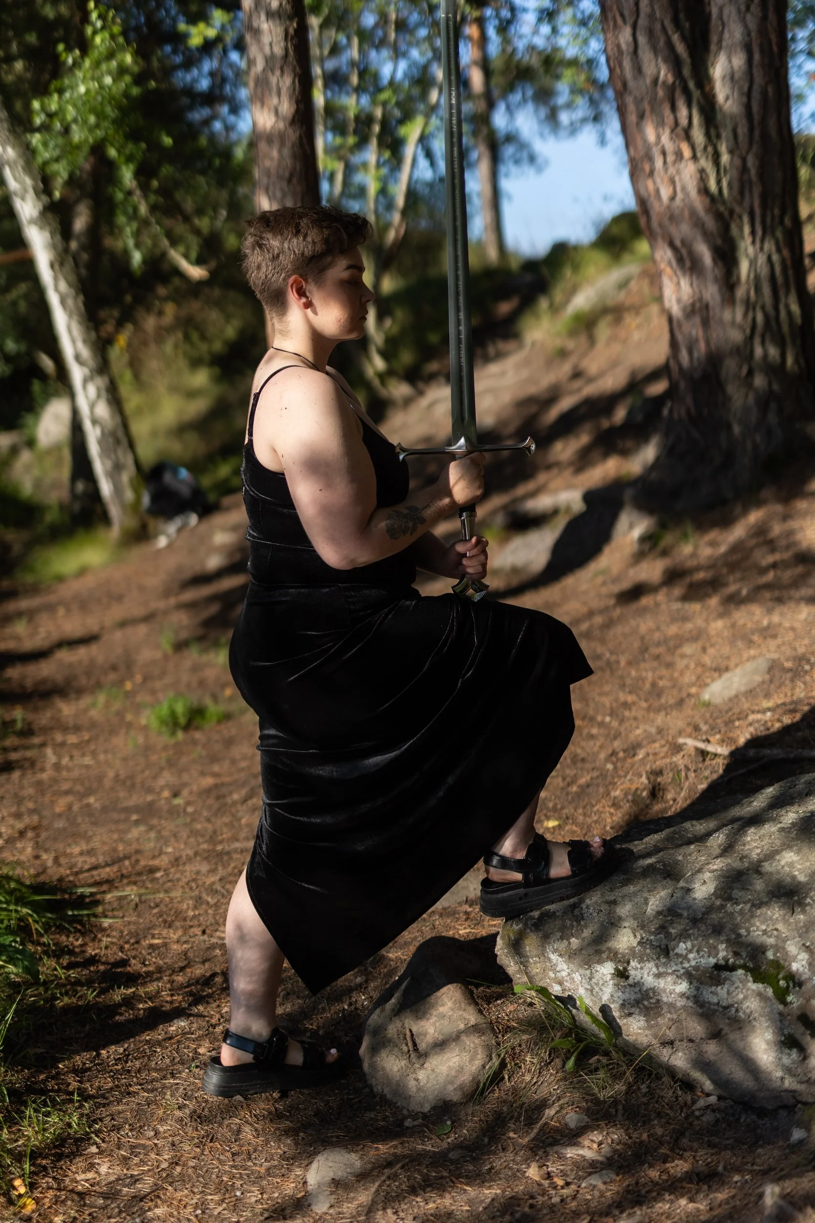 A person with short hair, wearing a black dress and sandals, kneels on the ground in a forest, holding a sword upright with both hands against their chest. They appear focused and serious, surrounded by tall trees and natural woodland scenery.