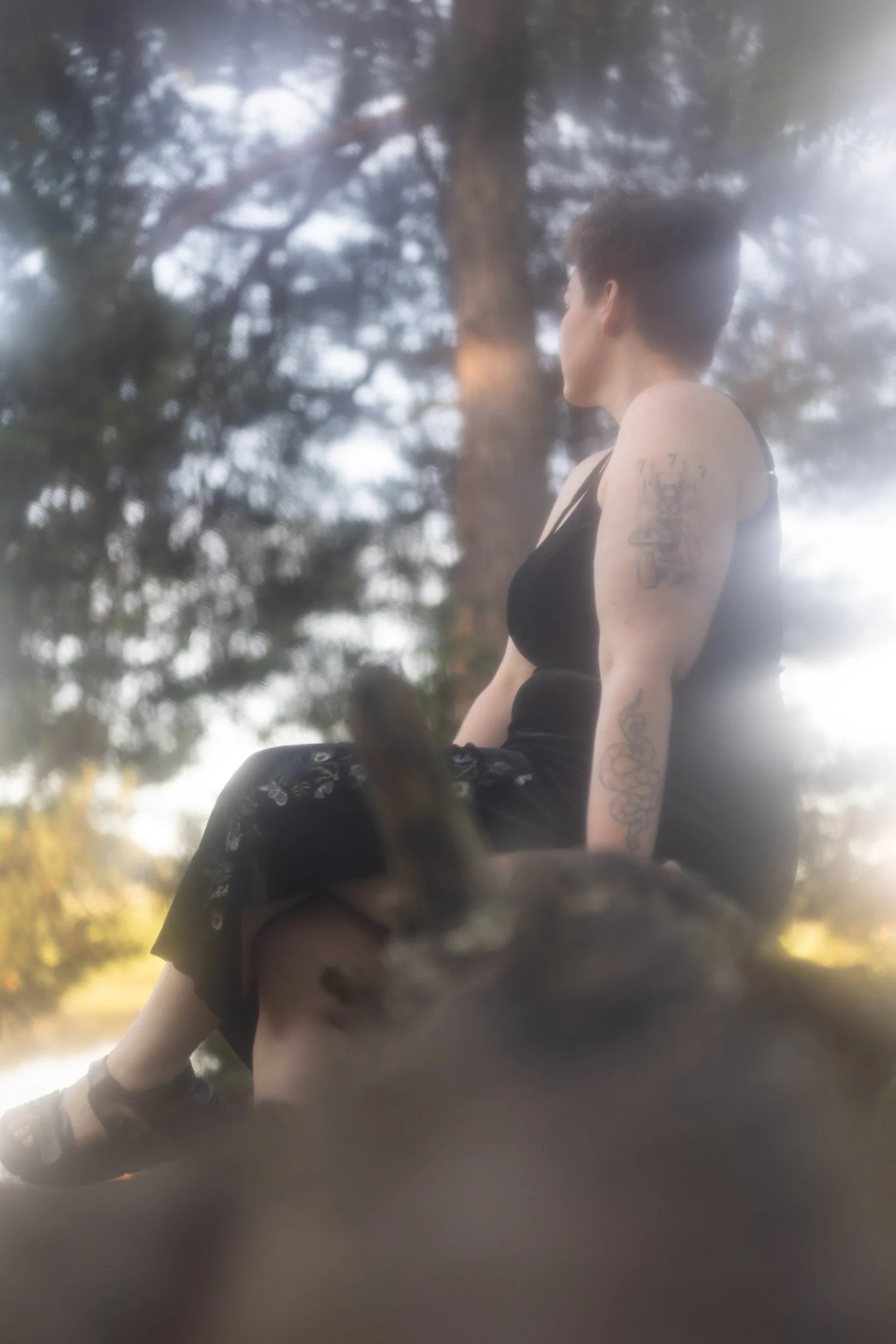A person with tattoos on their arm, sitting outdoors under trees, with a blurry background of green foliage. The person is facing to the right and is wearing a black tank top and dark pants, with a calm and contemplative expression.