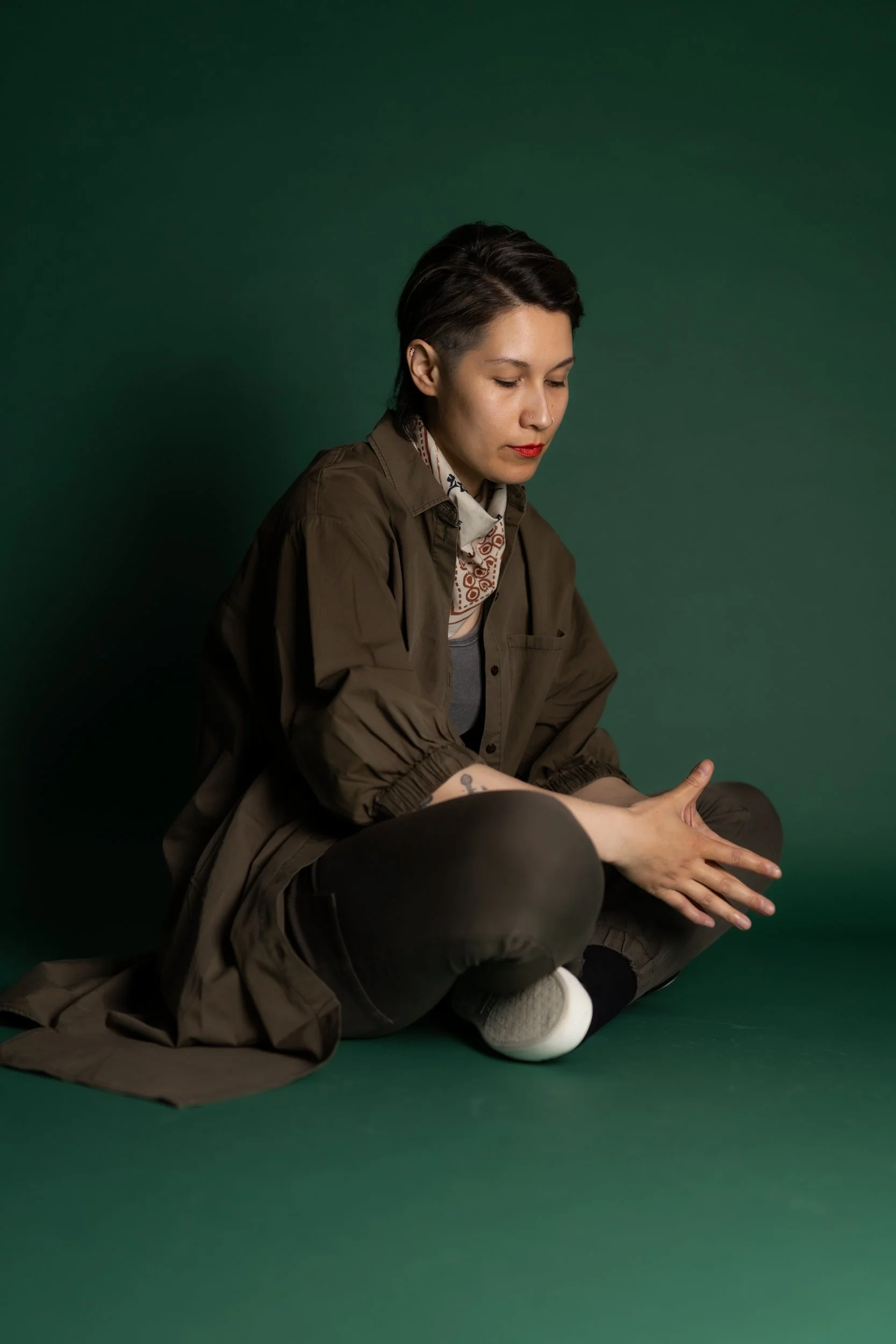 A person with short dark hair, wearing a brown trench coat and a patterned scarf, is sitting cross-legged against a solid green background, looking downward with their eyes closed and hands resting on their legs.