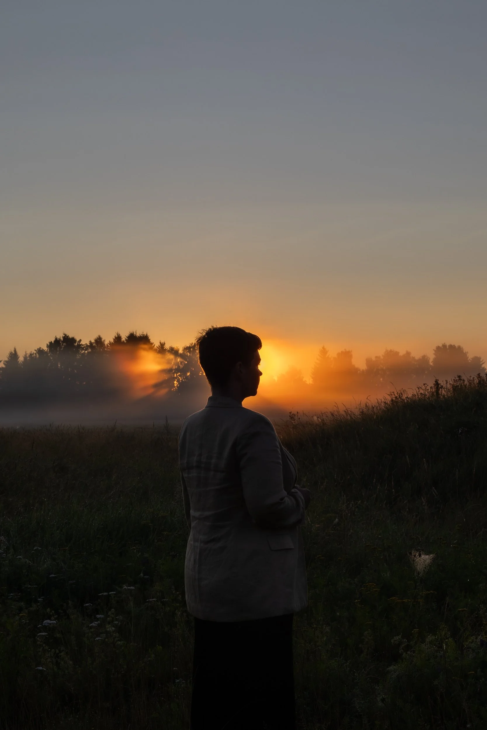 A person in a jacket stands outdoors during sunset with the sun setting behind a line of trees, creating a silhouette against the colorful sky.
