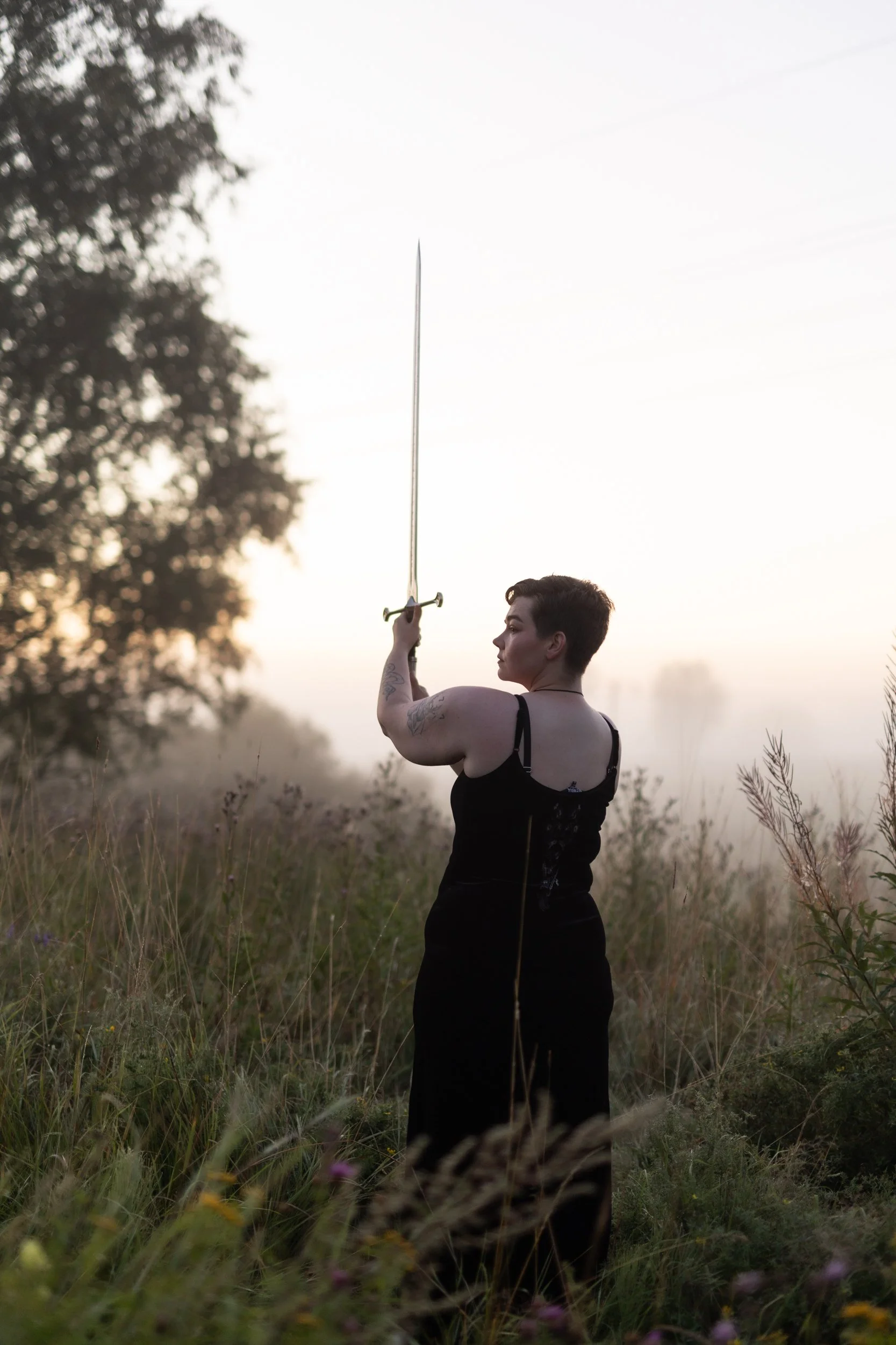 A person standing outdoors in a field at sunset, holding a sword upright above their head, with tall grass and wildflowers around them.