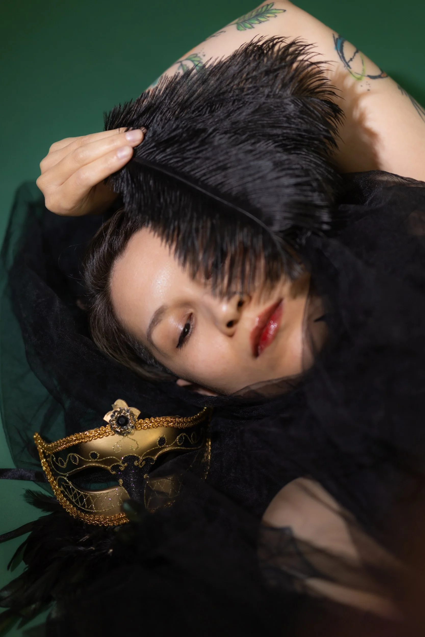 A person with short dark hair, lying down on a green surface, wearing gold jewelry and a black sheer garment, with tattoos on their arm.