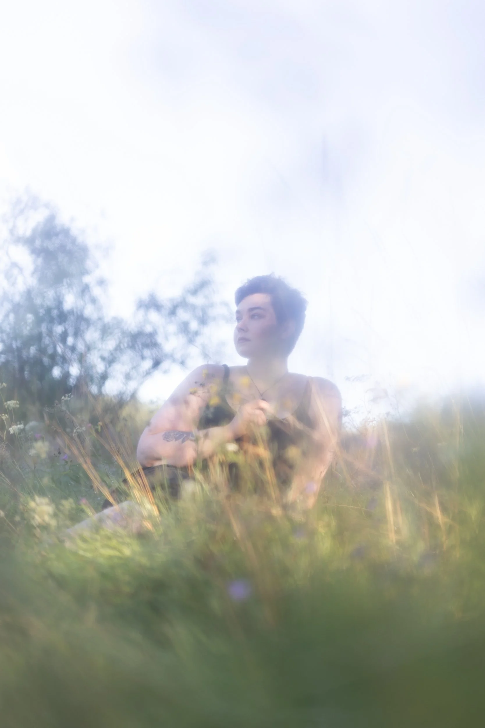 A person with short dark hair and a tattoo on their arm stands outdoors among tall grass and wildflowers, looking to the side with a thoughtful expression, with a bright sky and trees in the background.