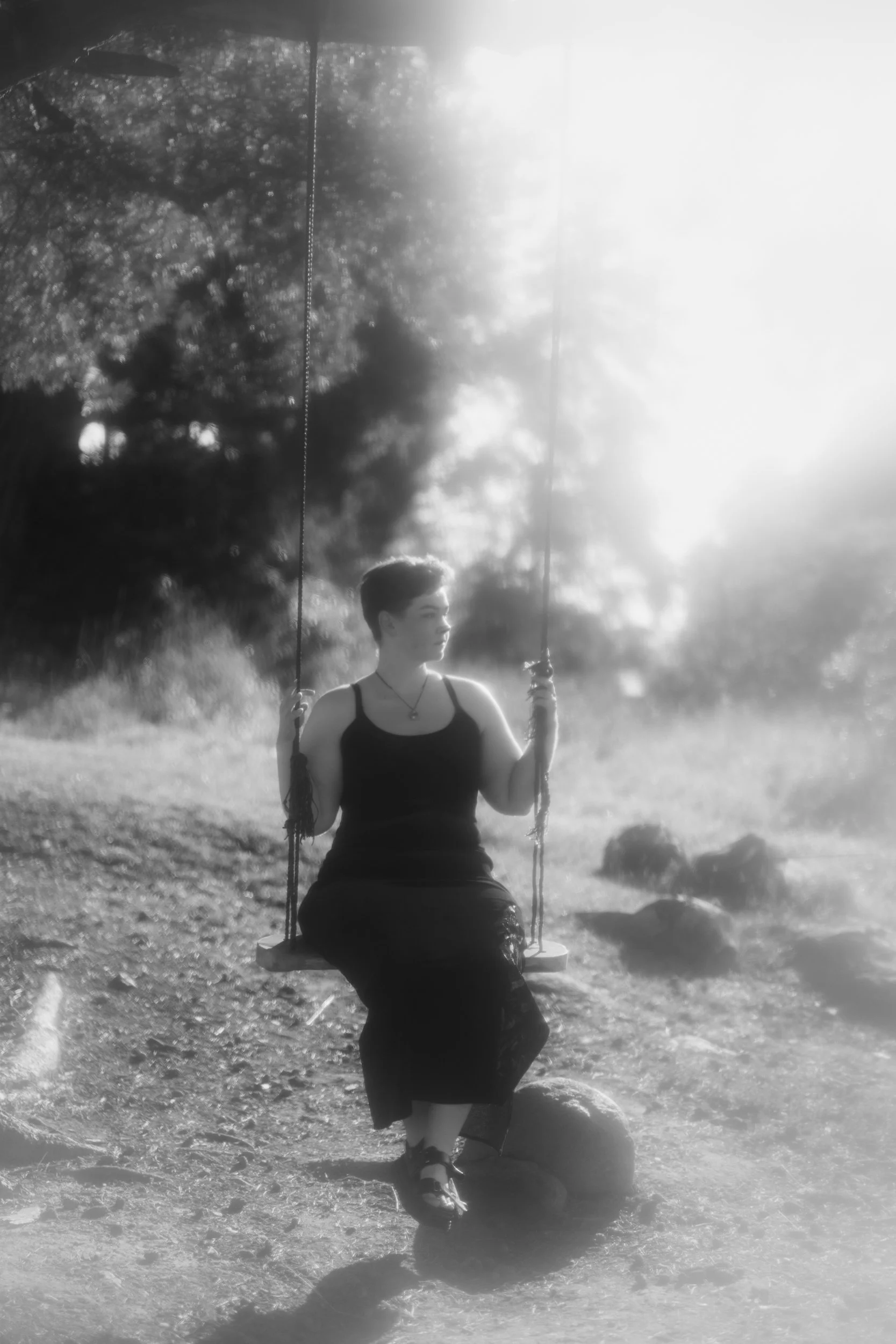 A person with short hair wearing a tank top and skirt sits on a swing in a natural outdoor setting with trees, rocks, and sunlight.