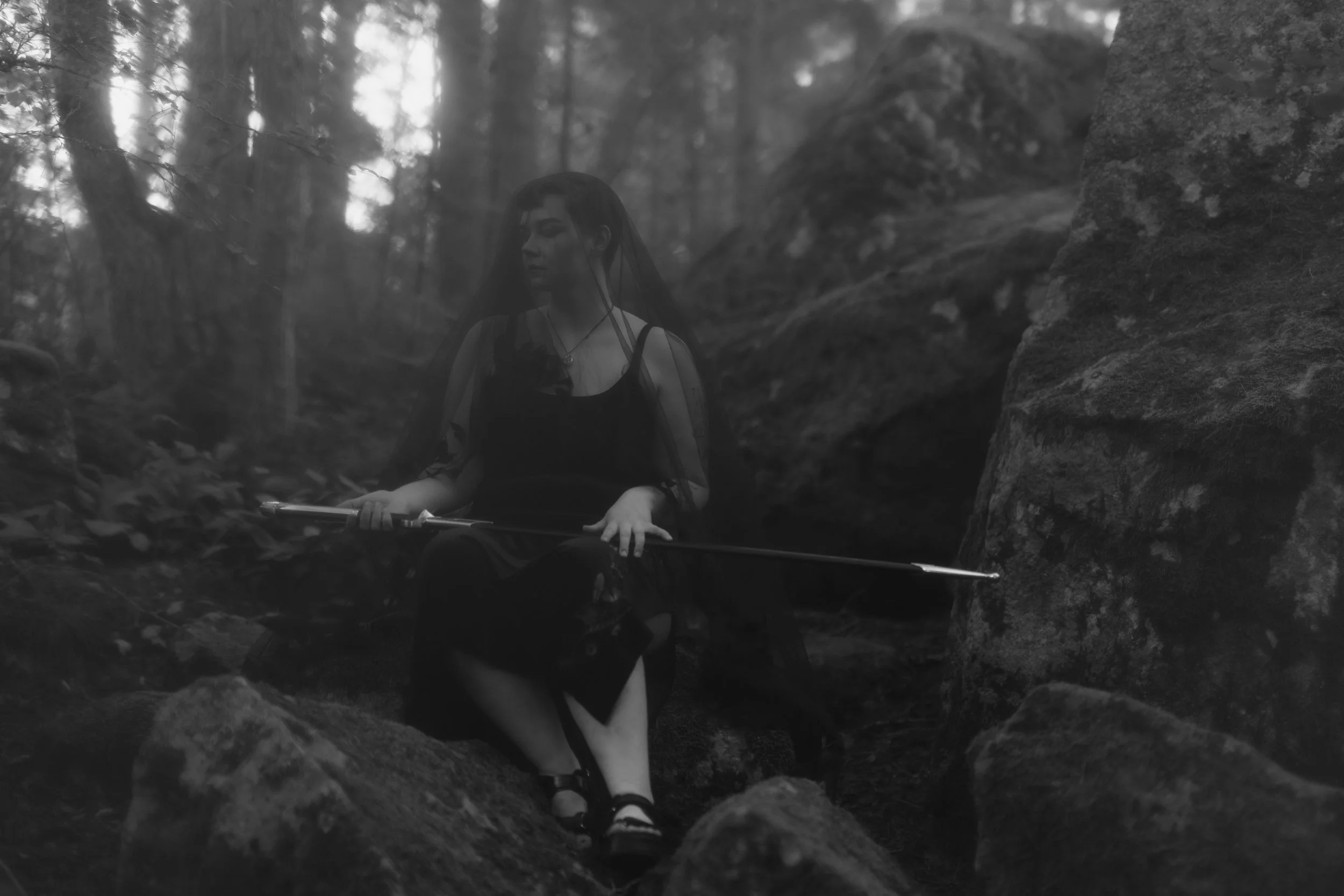 A person with dark hair and a veil, wearing a black dress, is sitting on a rock in a forested area with large rocks and trees around them. They are holding a sword on their lap. The image is in black and white.