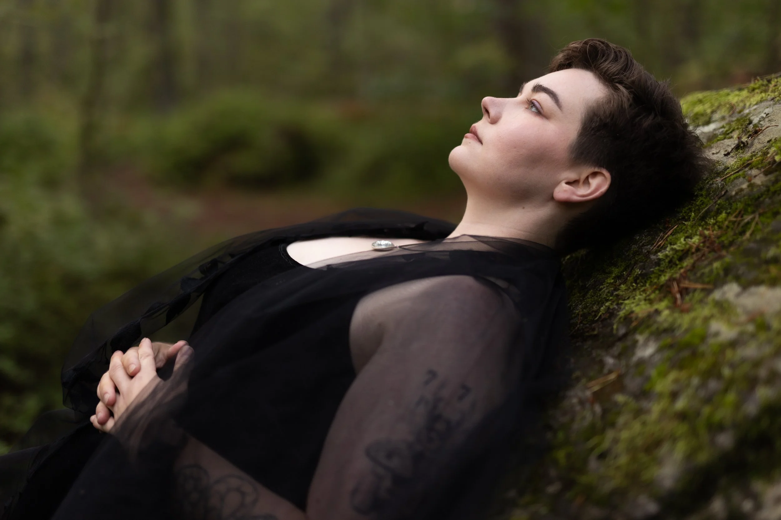 A person with short curly hair lying on their back on the ground in a forest, eyes closed, hands clasped together over their stomach.