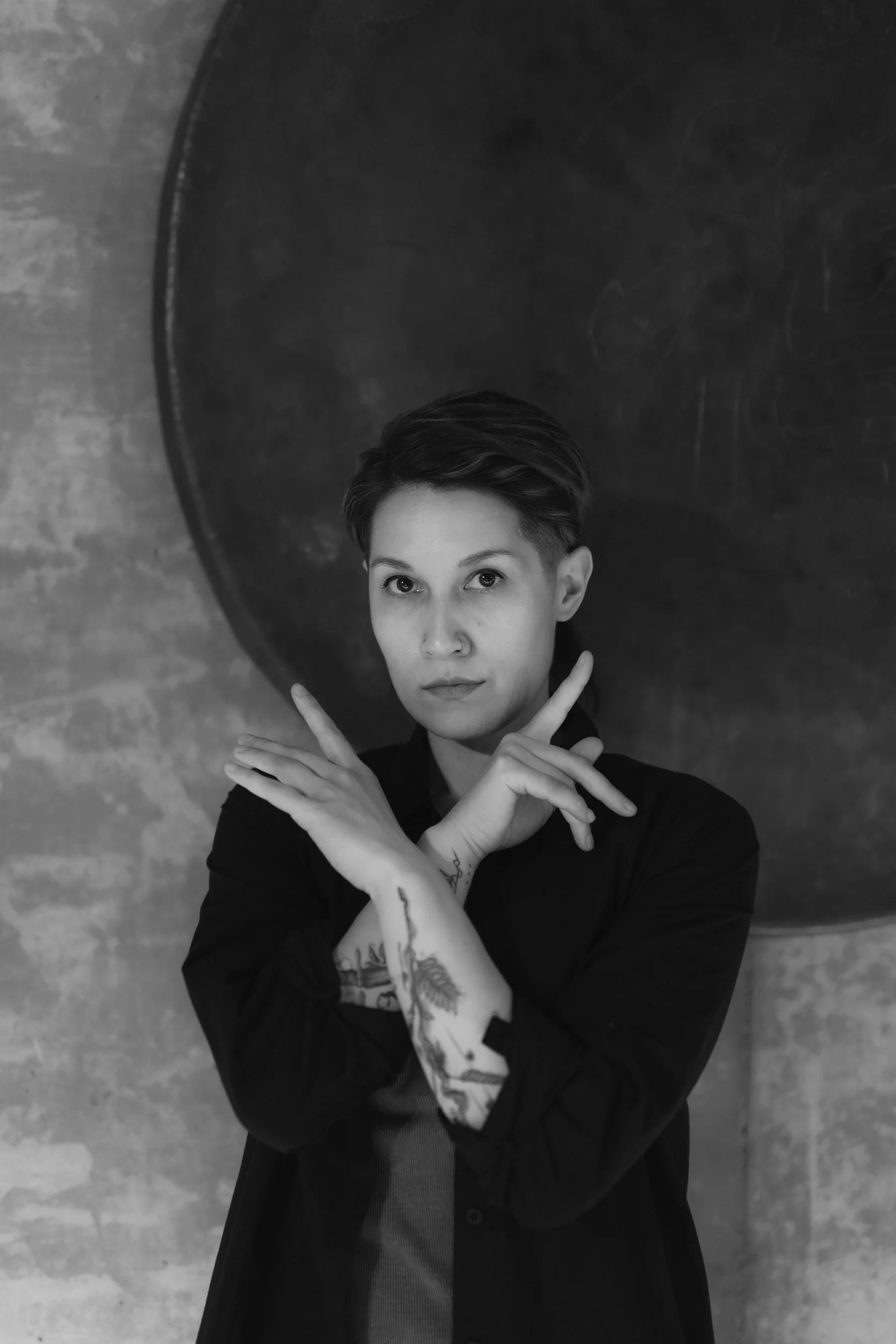 A black and white photo of a person with short, styled hair making an X shape with their arms crossed in front of their chest. The person has a nose ring and visible tattoos on their arms, and is wearing a dark top. The background consists of a textu