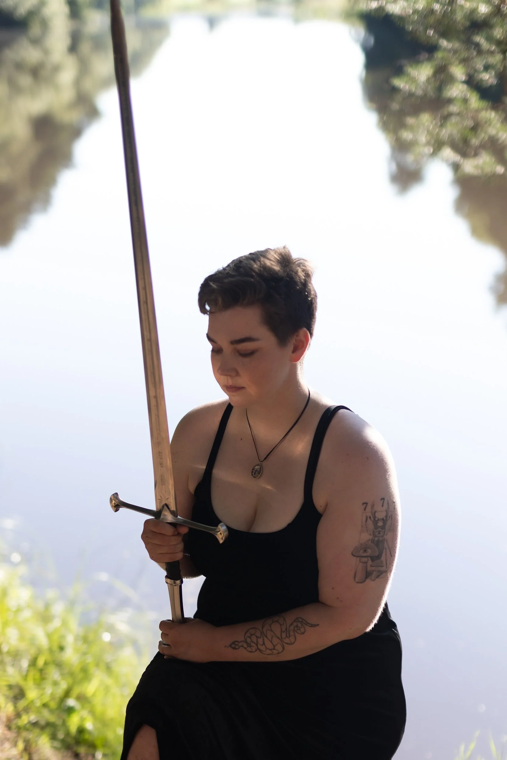 A person with short dark hair, tattoos on her arm, and wearing a black tank top holds a sword while standing outdoors near a body of water, surrounded by trees.