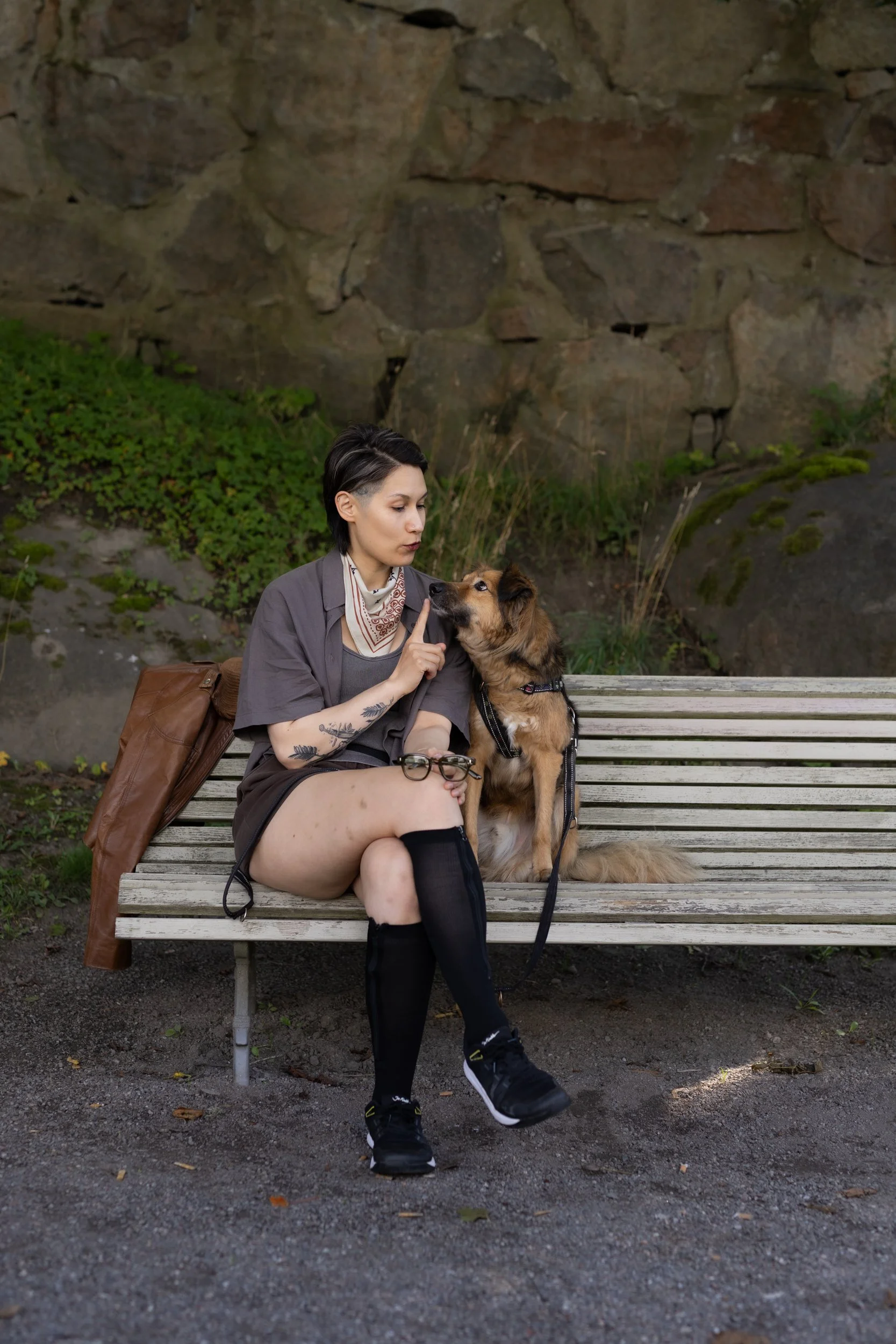 A person with short dark hair and tattoos sitting on a park bench with a brown and black dog, against a background of a stone wall and greenery.