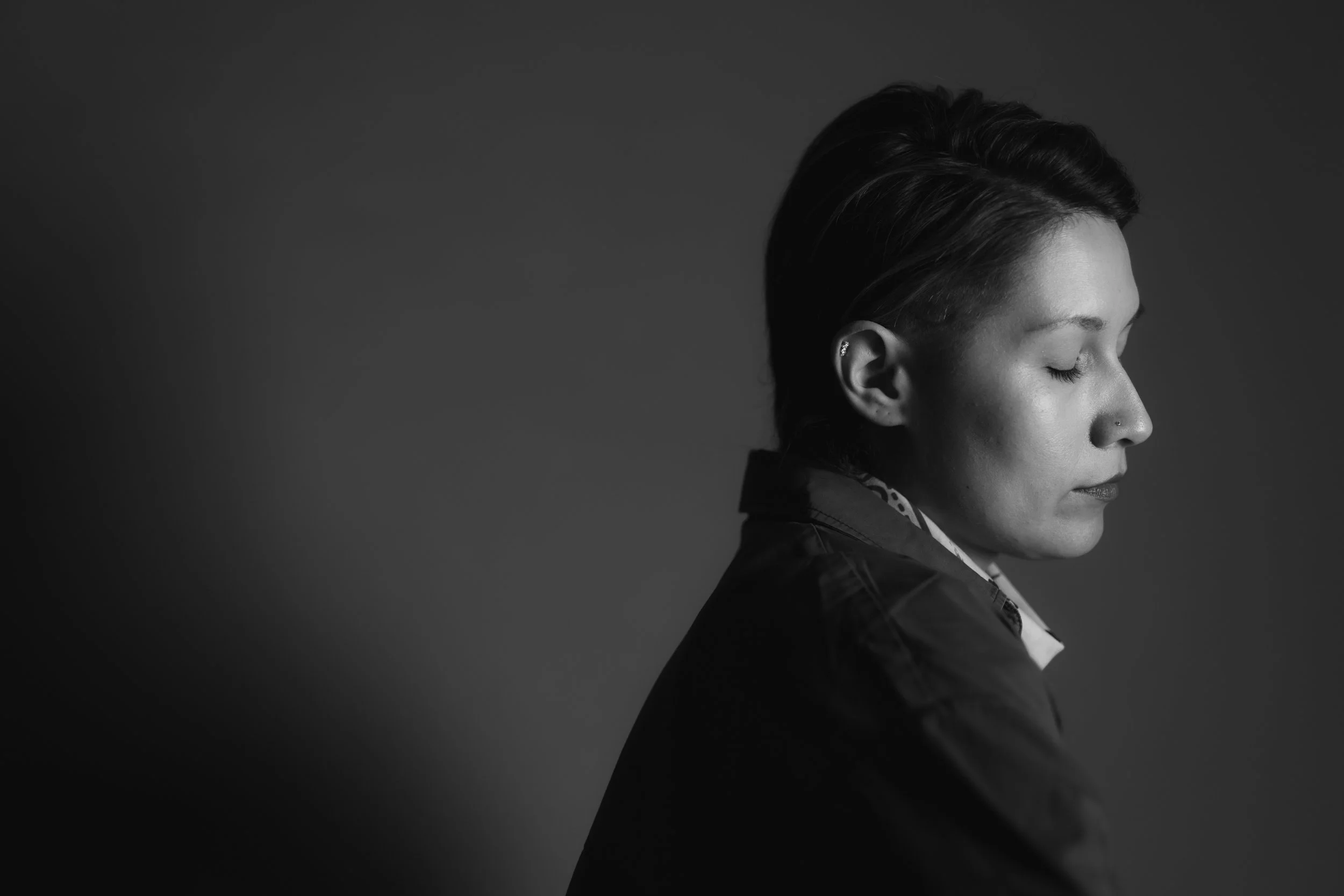 A black and white portrait of a person with short dark hair, eyes closed, wearing a jacket, and a small earring in their left ear.