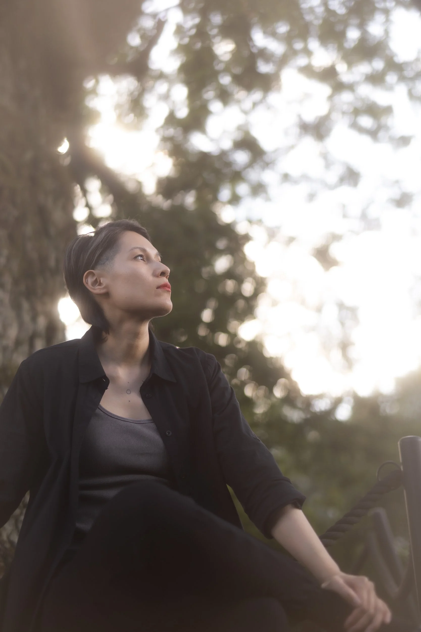 A person with short dark hair, wearing a black jacket over a gray top, sitting outdoors, looking to their left with a serene expression. The background features trees and sunlight filtering through the leaves.