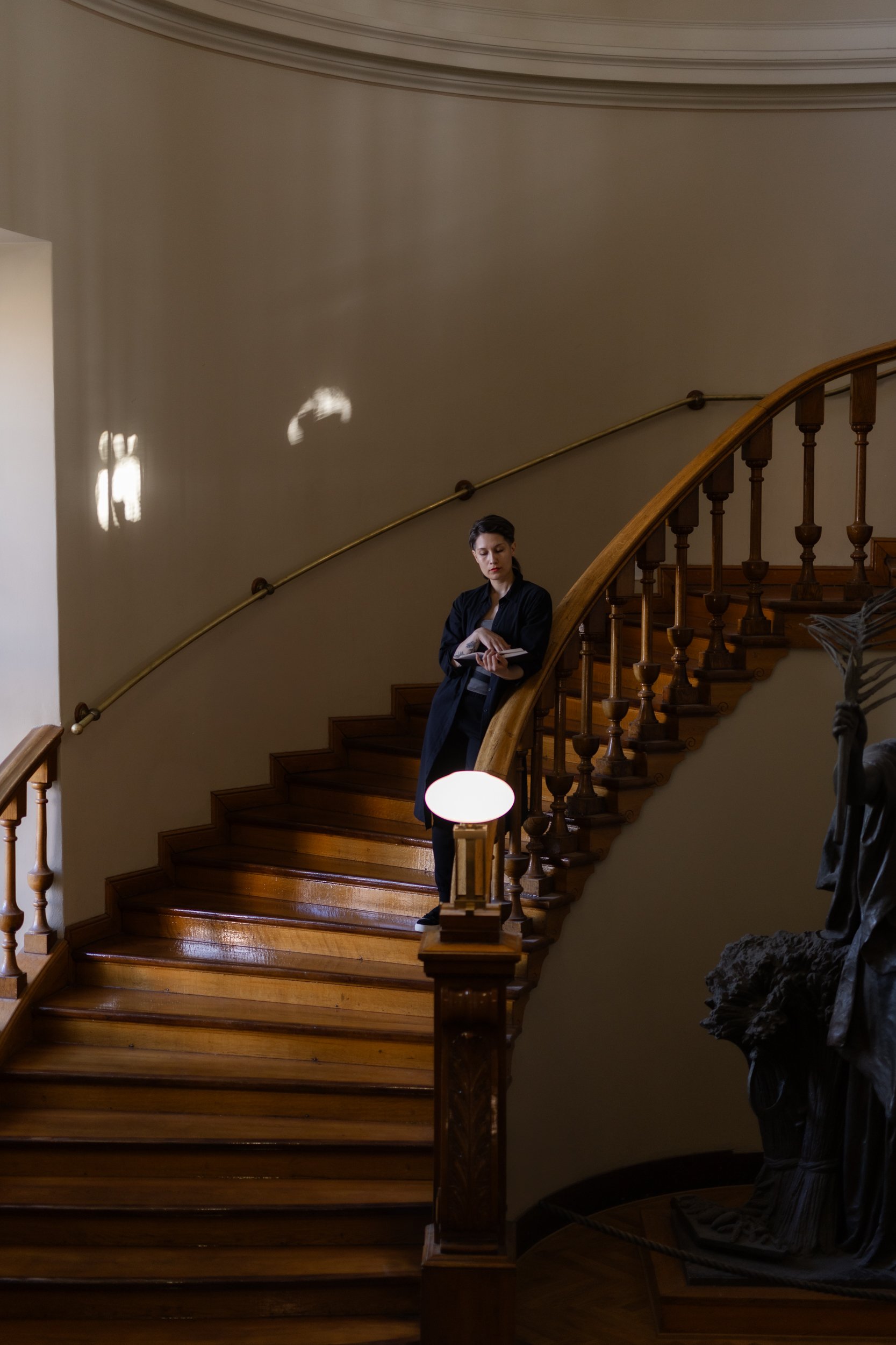 A person with dark hair, wearing a black jacket, stands on a wooden staircase, holding a notebook, with a wooden railing and a bronze statue of a man nearby, in a warmly lit interior.