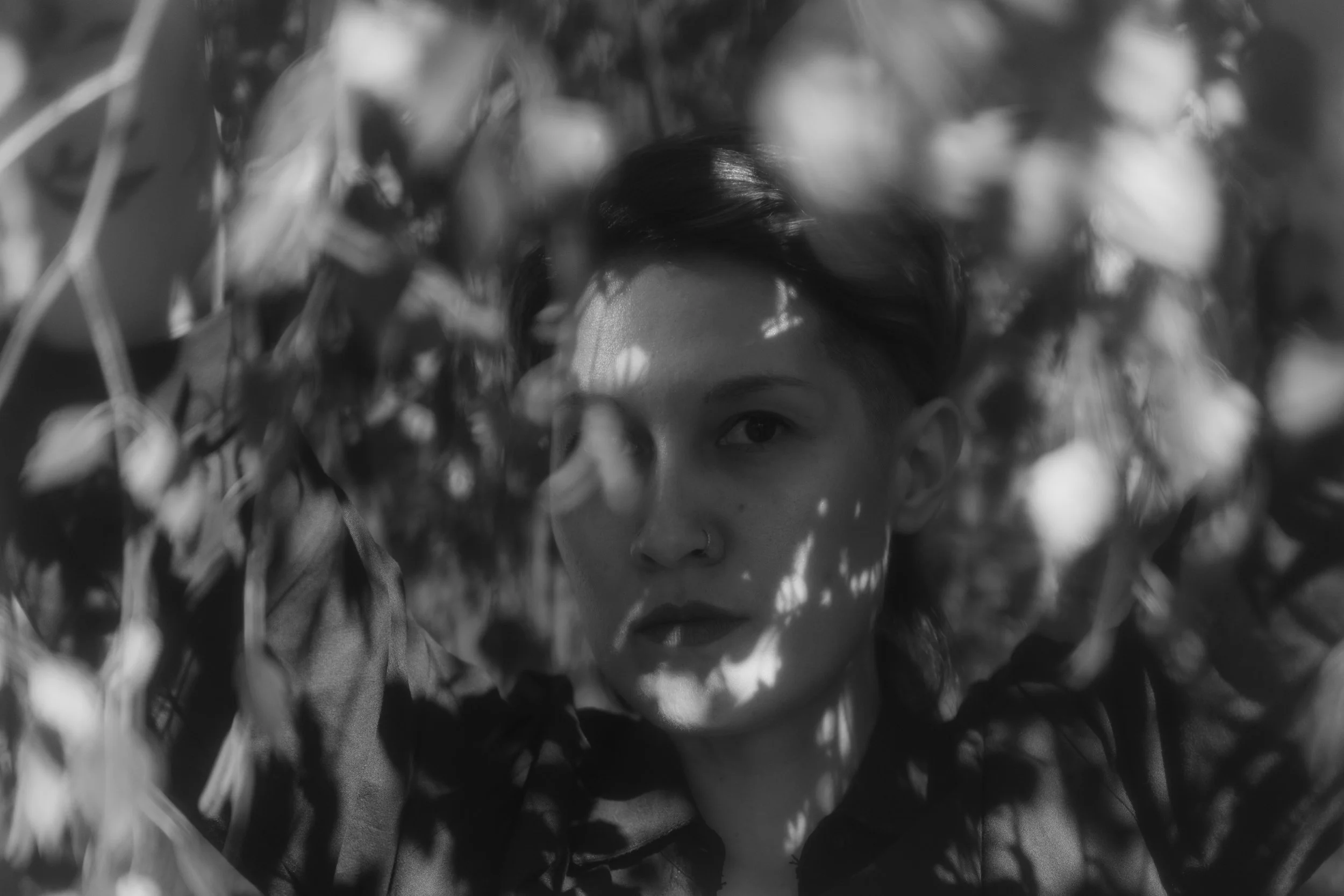 Black and white close-up photo of a person's face partially hidden among blurred leaves and branches.