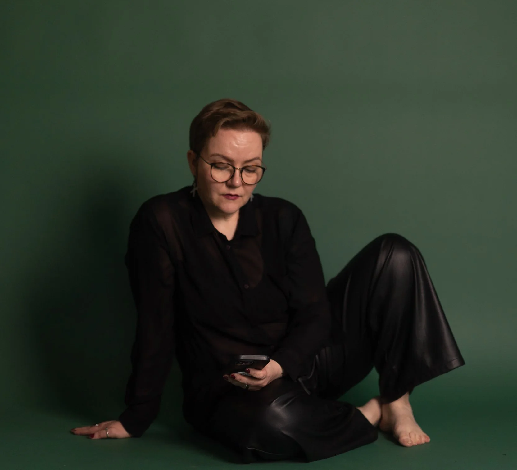 Photographer sit in front of a green back drop. They have a phone on their hand and theey look down at it. They are wearing a black mesh blouse and black leather pants. They do not have shoees on.