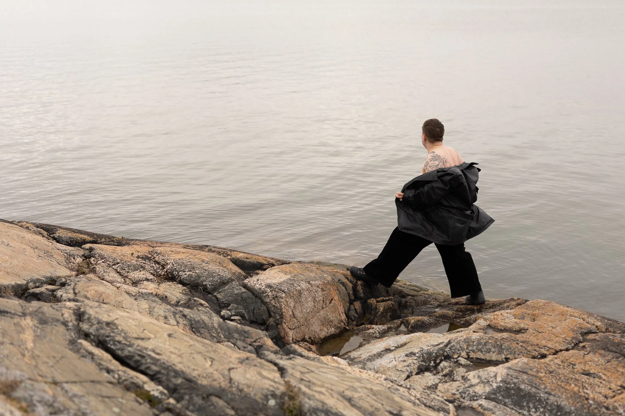 Portrait of Siiri walking on the rocky shoreline at Ruissalo in Turku, wearing a leather jacket pulled off her shoulders so her bare shoulders are visible, photographed from behind.