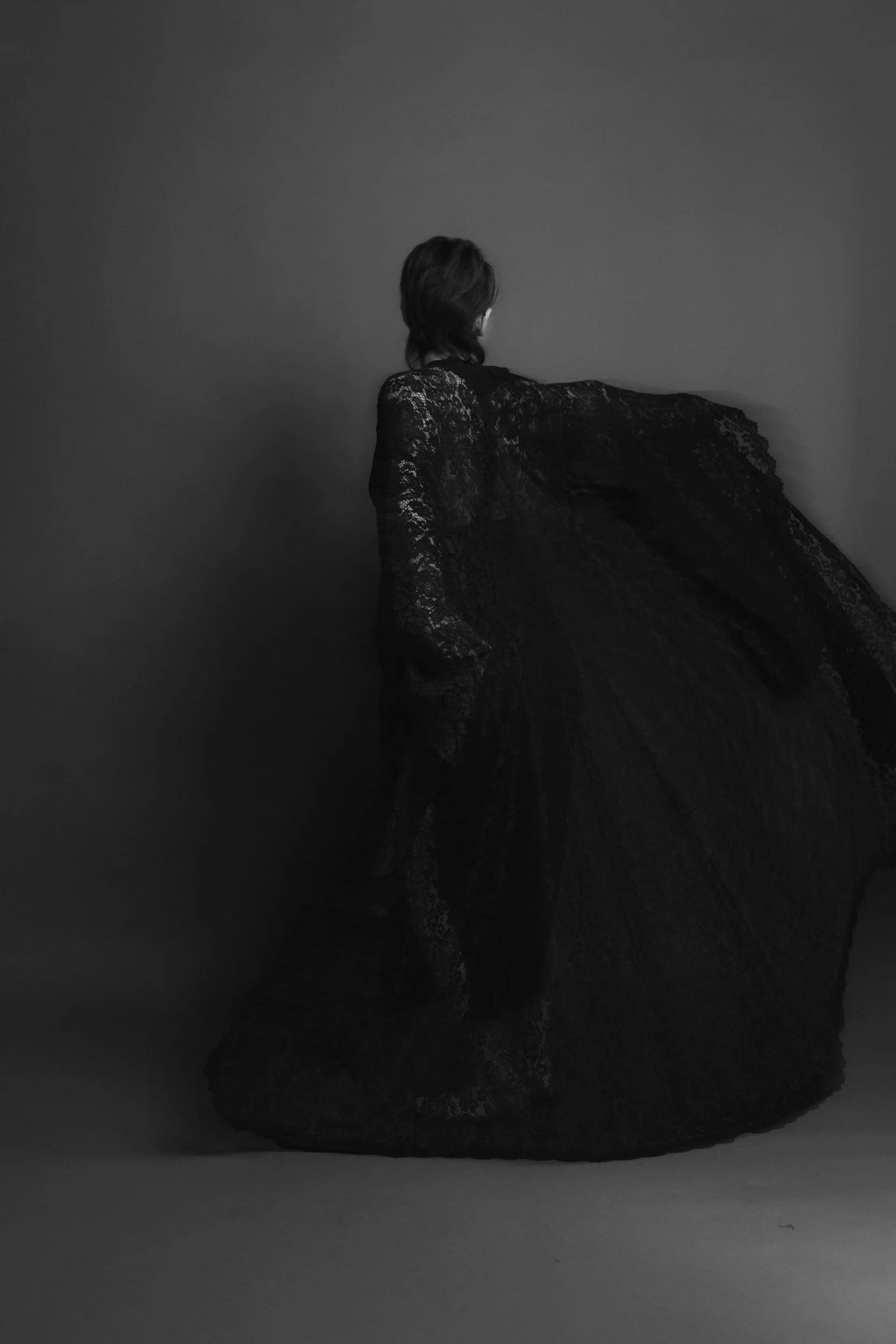 Black-and-white photo of a person in lace dress, standing sideways with their head turned away, against a plain background.