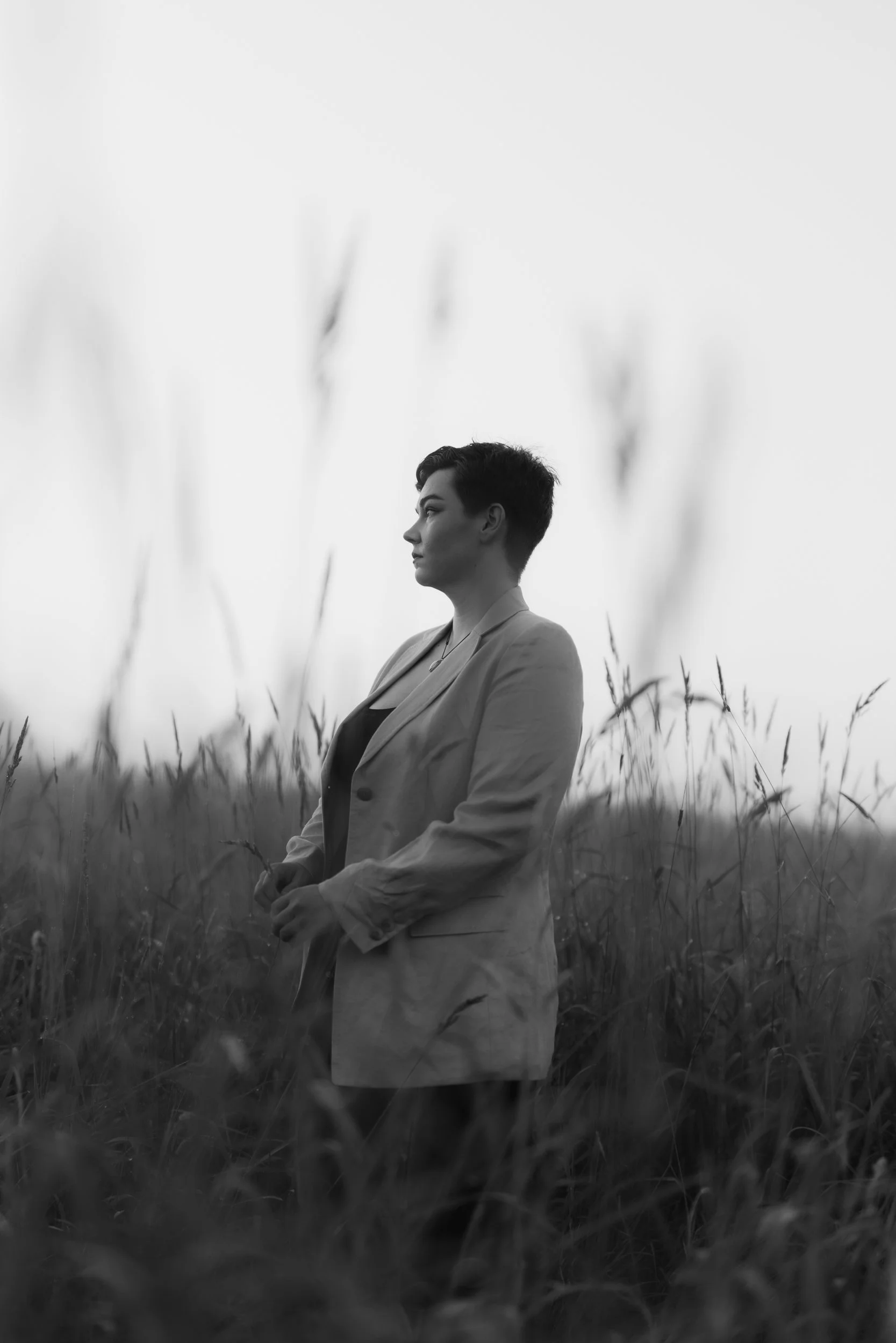 Black and white photo of a person in a jacket standing in a field of tall grass, looking to the side with a contemplative expression.
