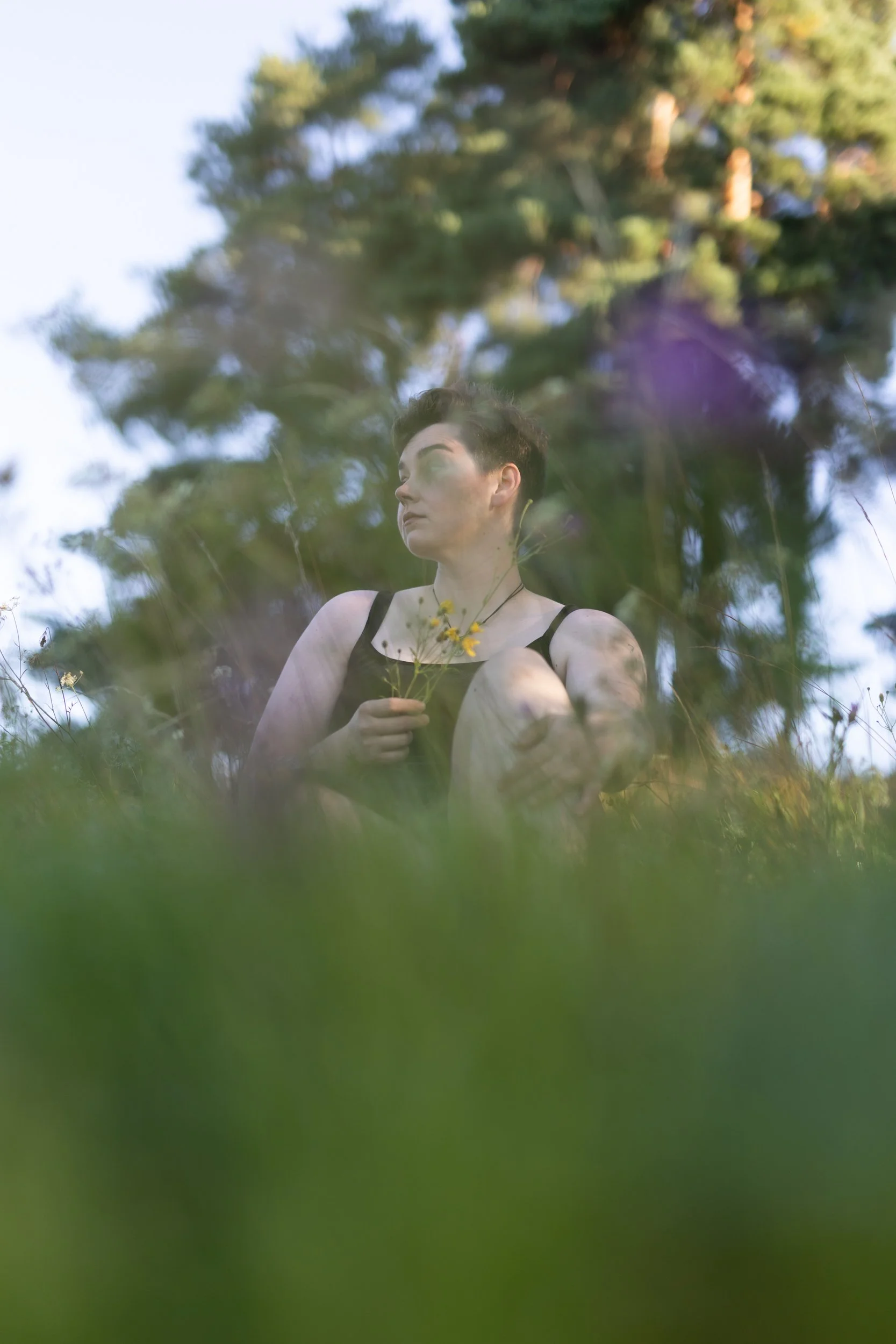 A person with short dark hair and a black tank top is sitting outdoors among trees, holding a small bunch of yellow flowers, with a blurred green foreground and trees in the background.