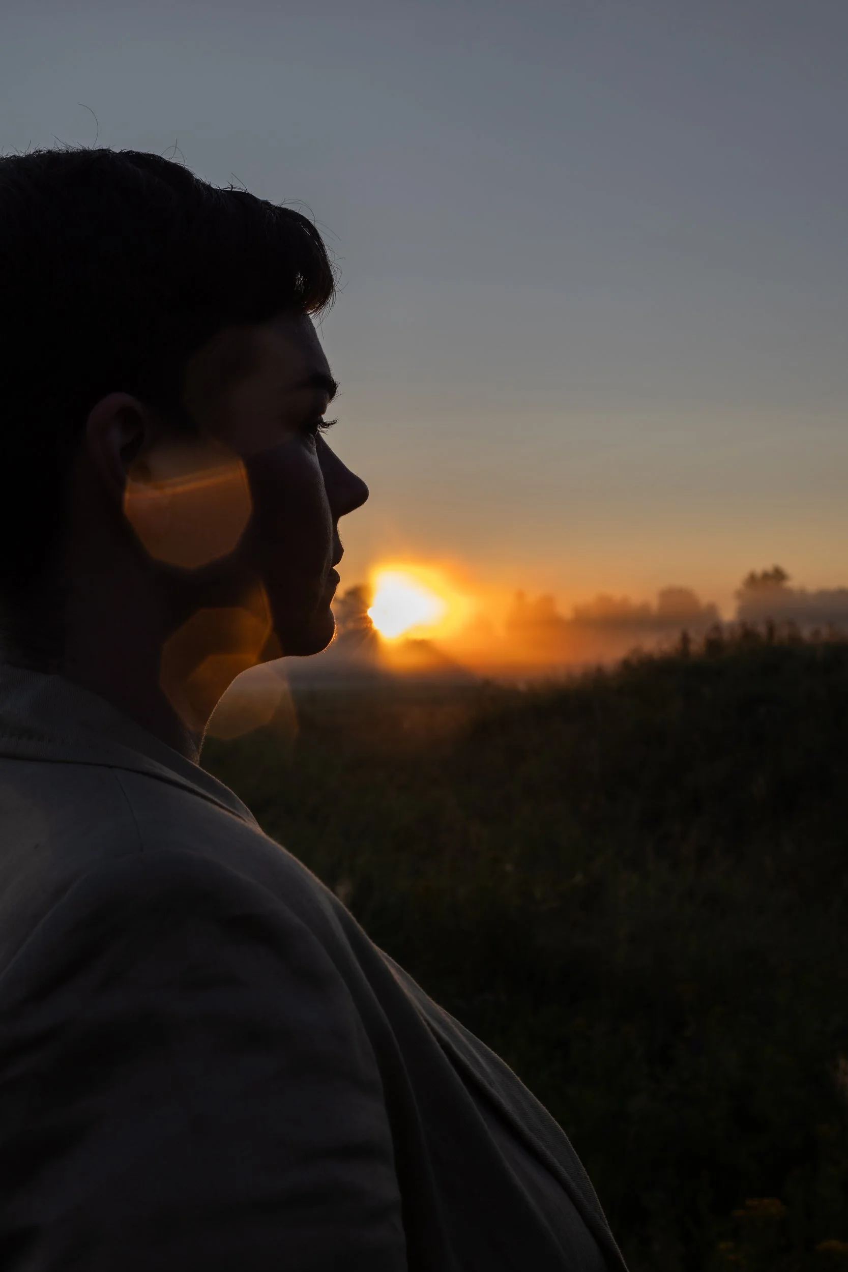 Silhouette of a person with short hair wearing a hoodie, facing sunset with a partly cloudy sky and distant horizon.