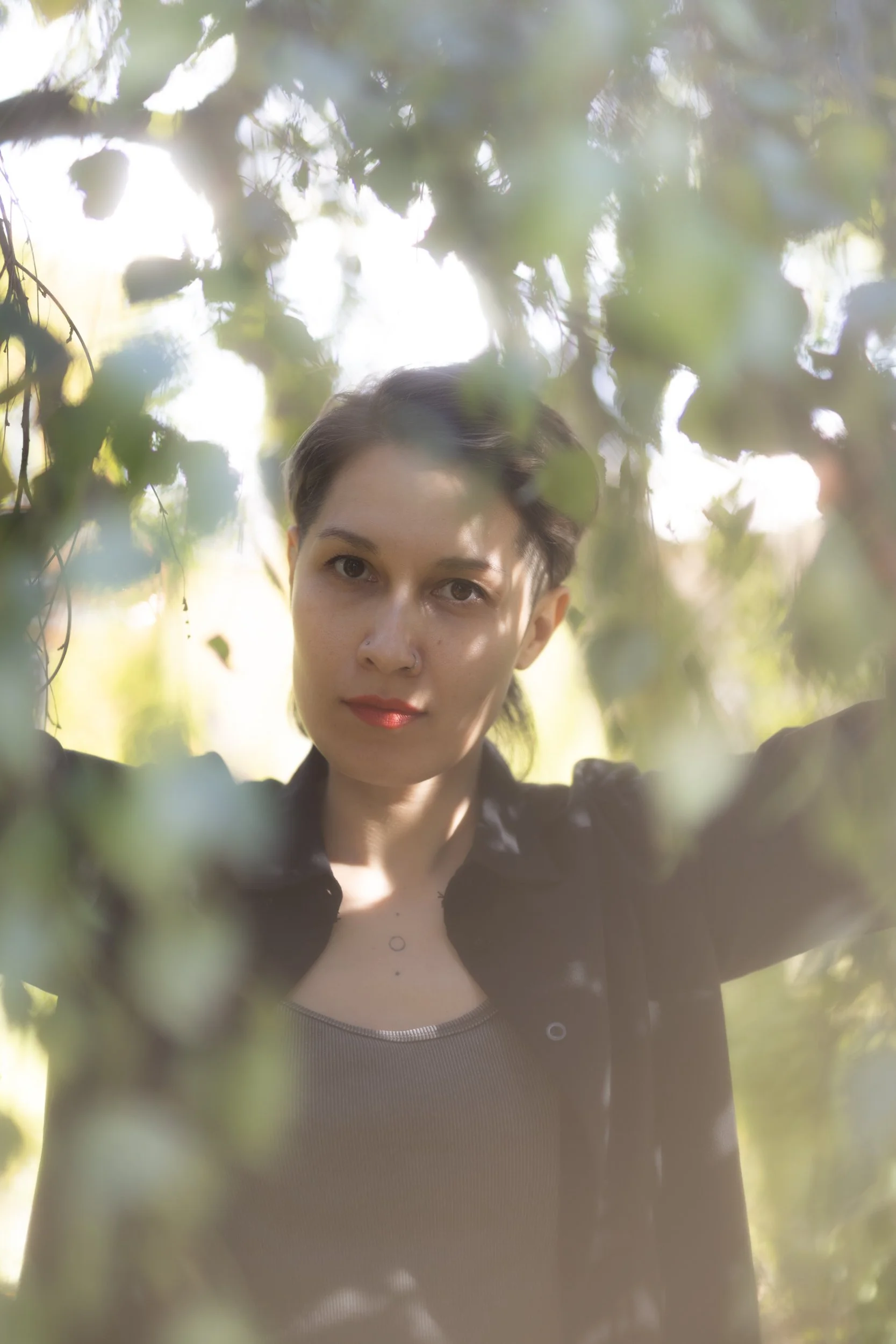 A person with short dark hair and a nose ring, wearing a black jacket and a gray shirt, looking through blurred green leaves with sunlight filtering through.