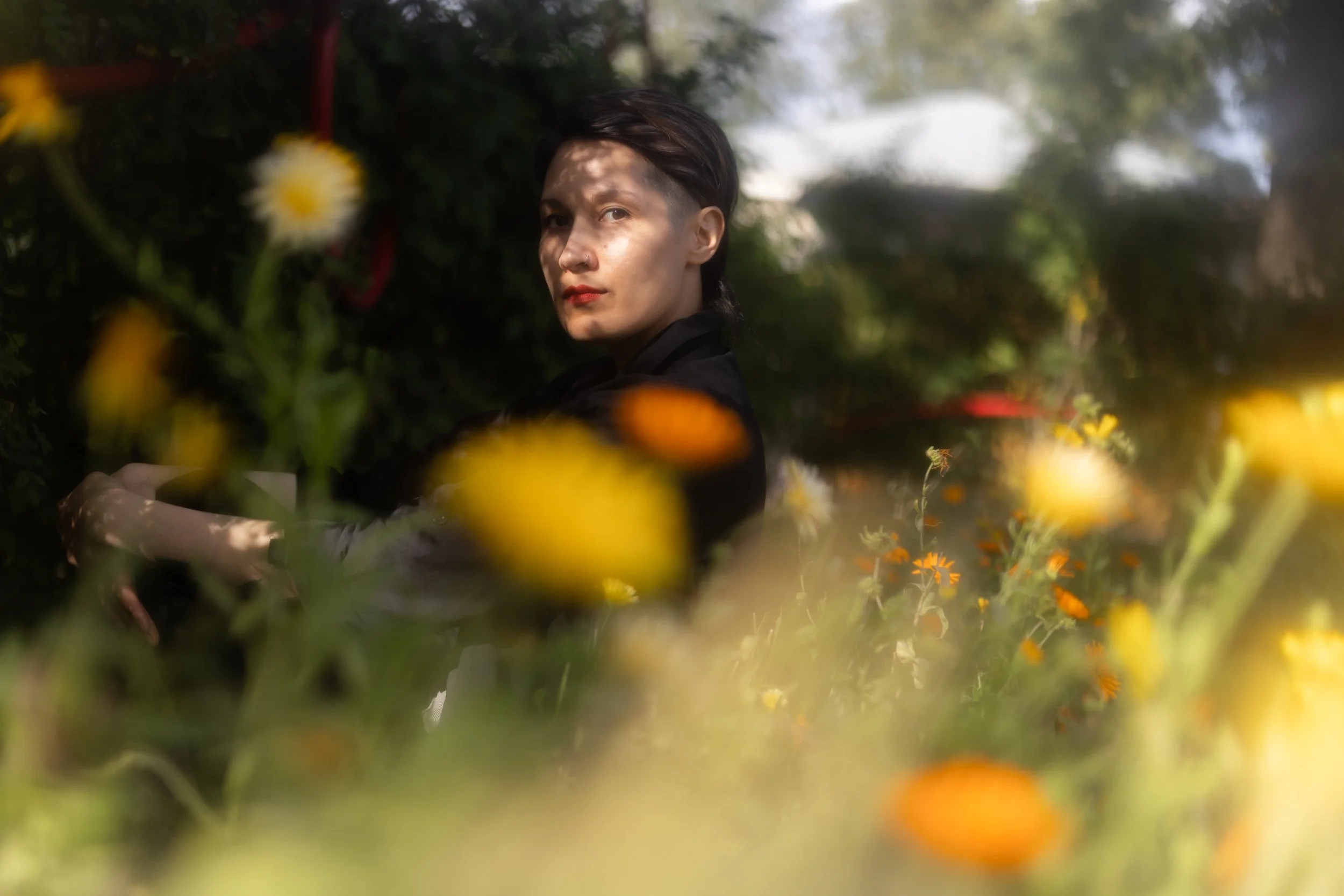 A person with short dark hair and wearing dark clothing among greenery and orange flowers, looking at the camera.
