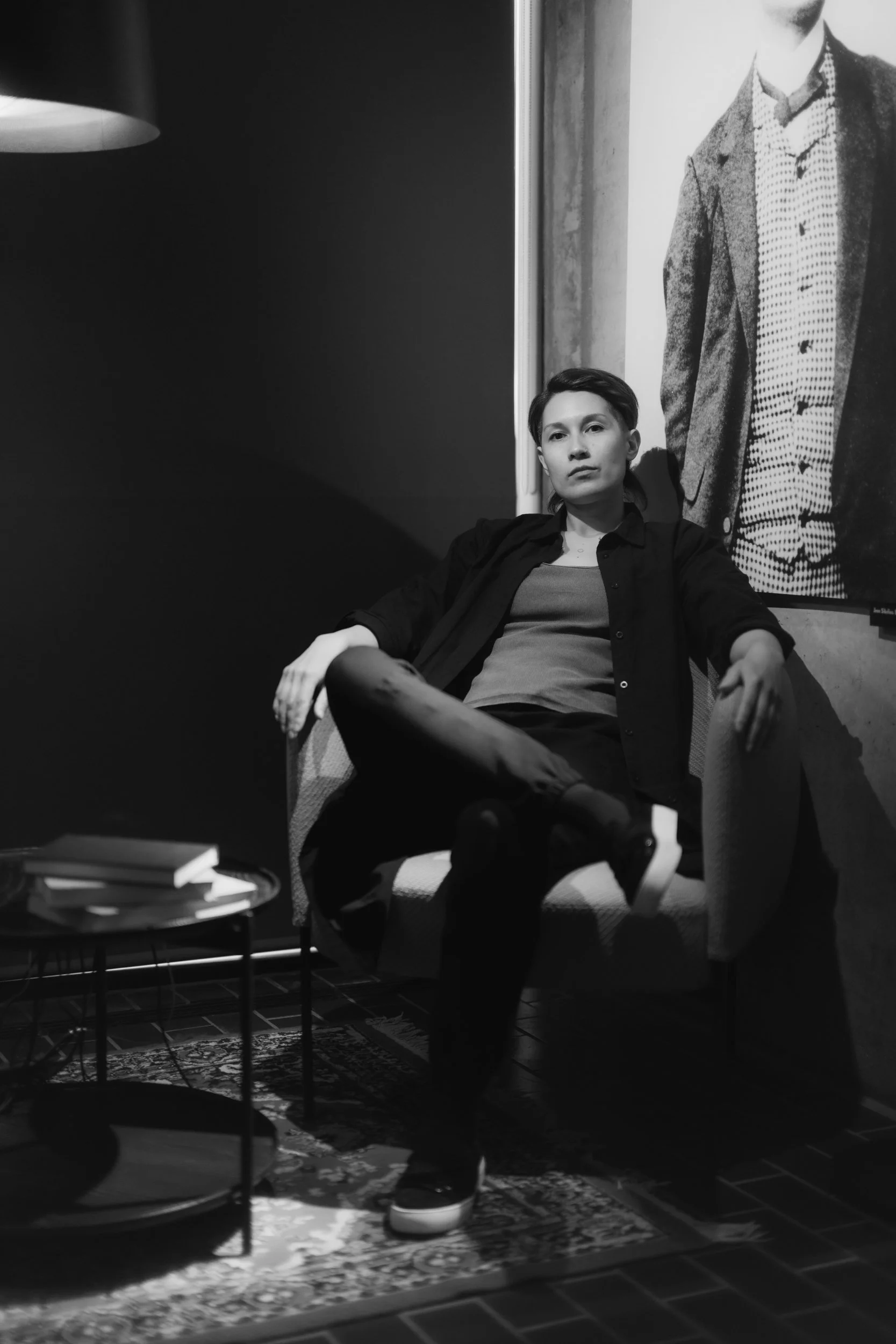 A young woman with short hair sitting in a chair in a black-and-white photo. Behind her, a large portrait of a man in a blazer and polka dot shirt is visible on the wall. There is a small table with books and a patterned rug on the floor.