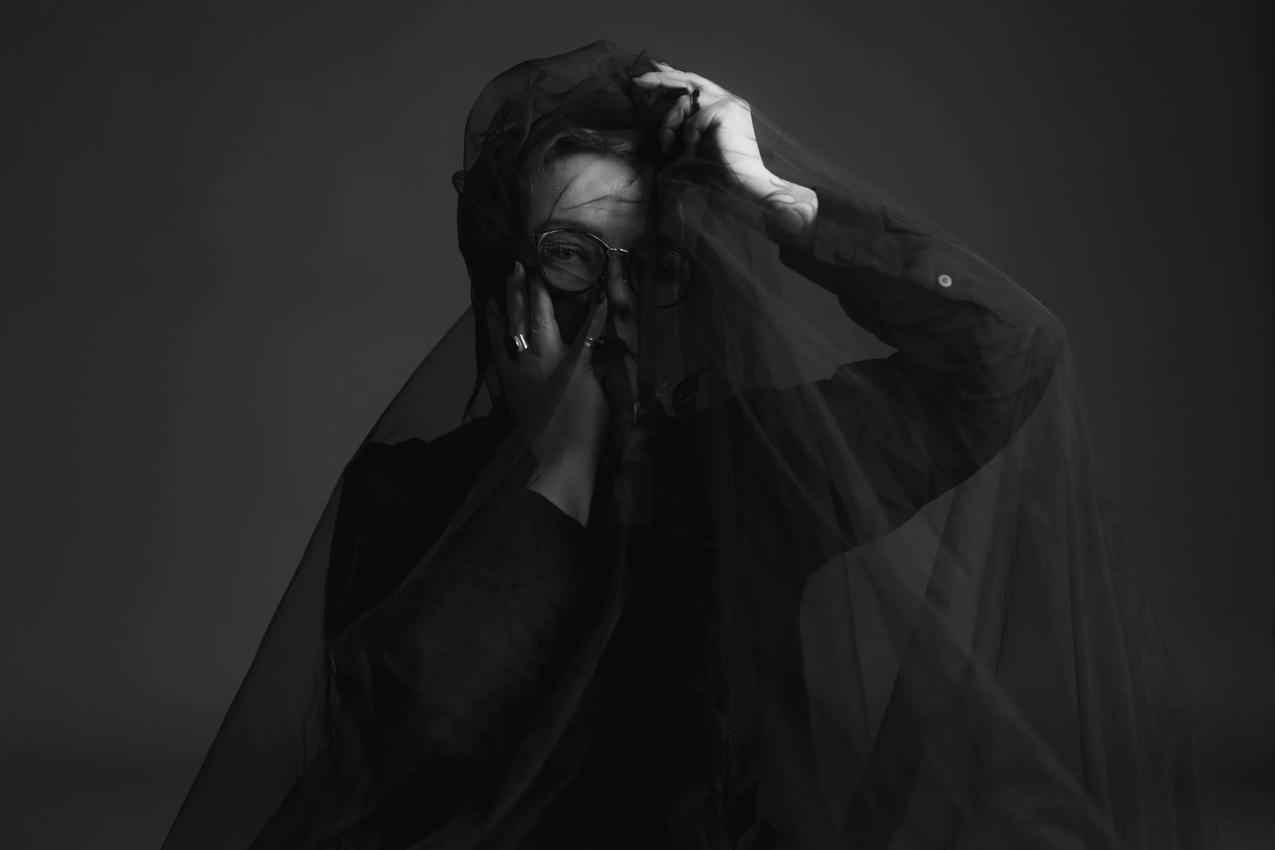 Tinksu is facing the camera. They are under a black veil and their hands are near their face.