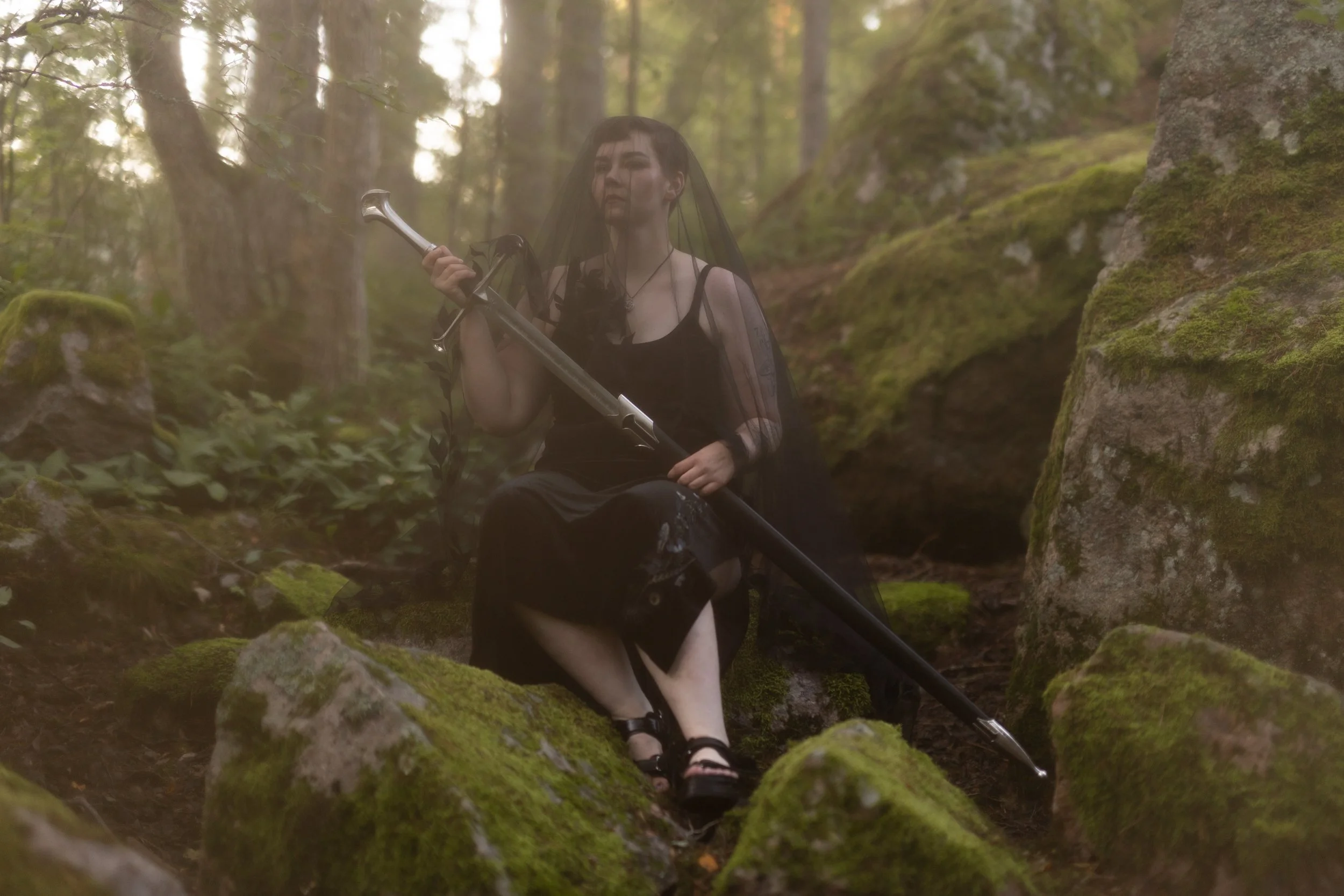 A person in black attire holding a sword, sitting on moss-covered rocks in a lush forest with tall trees and sunlight filtering through the leaves, veiled in a sheer black fabric.