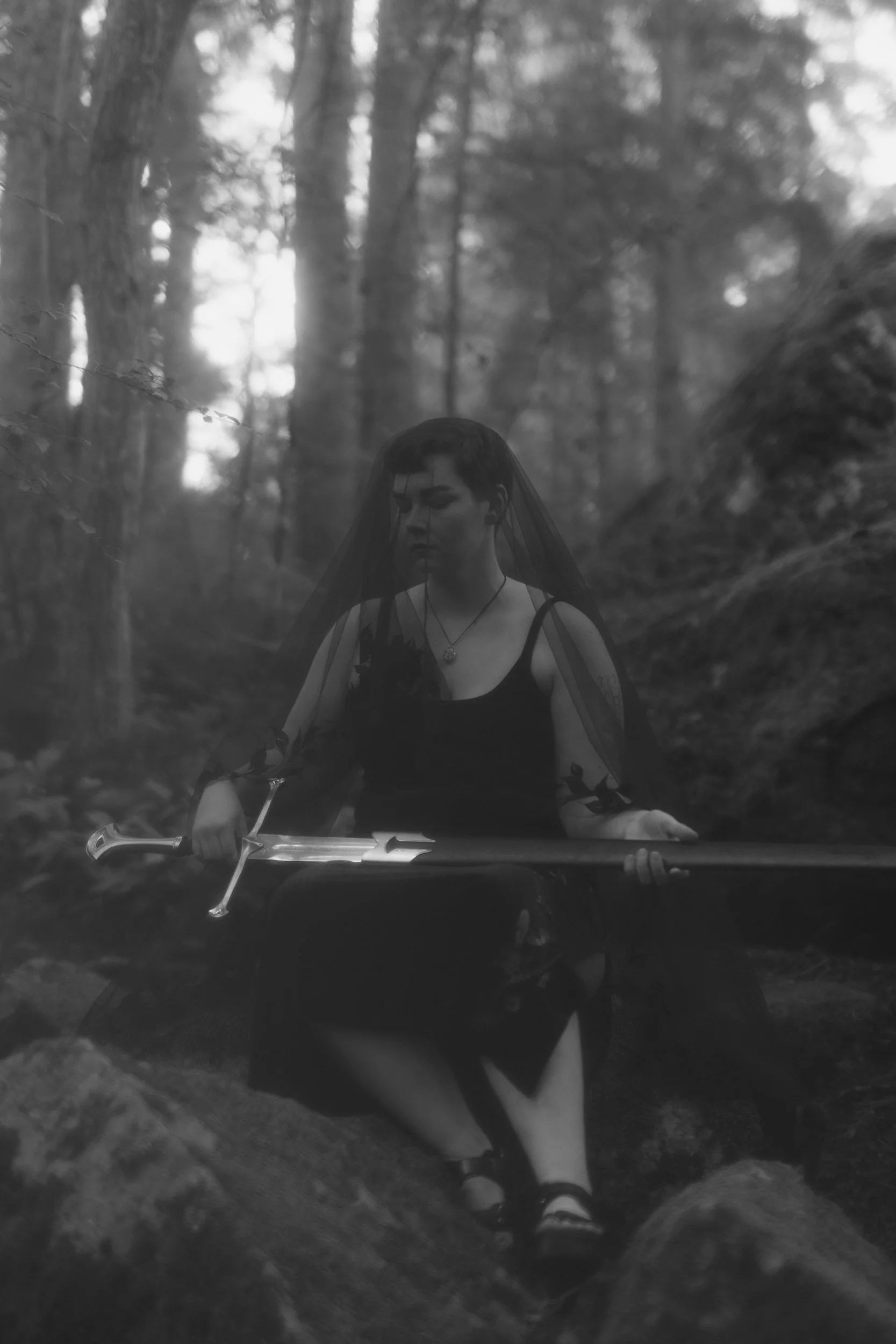 A person sitting on a rock and holding a sword in a forest, wearing a veil and casual clothing, with trees and soft background lighting in black and white.