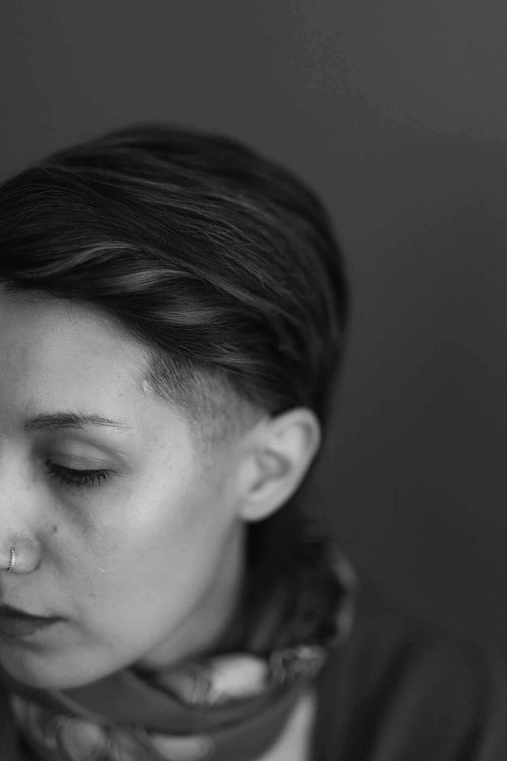 Close-up of a person's face with closed eyes, gray colored hair, and a nose piercing, in black and white.