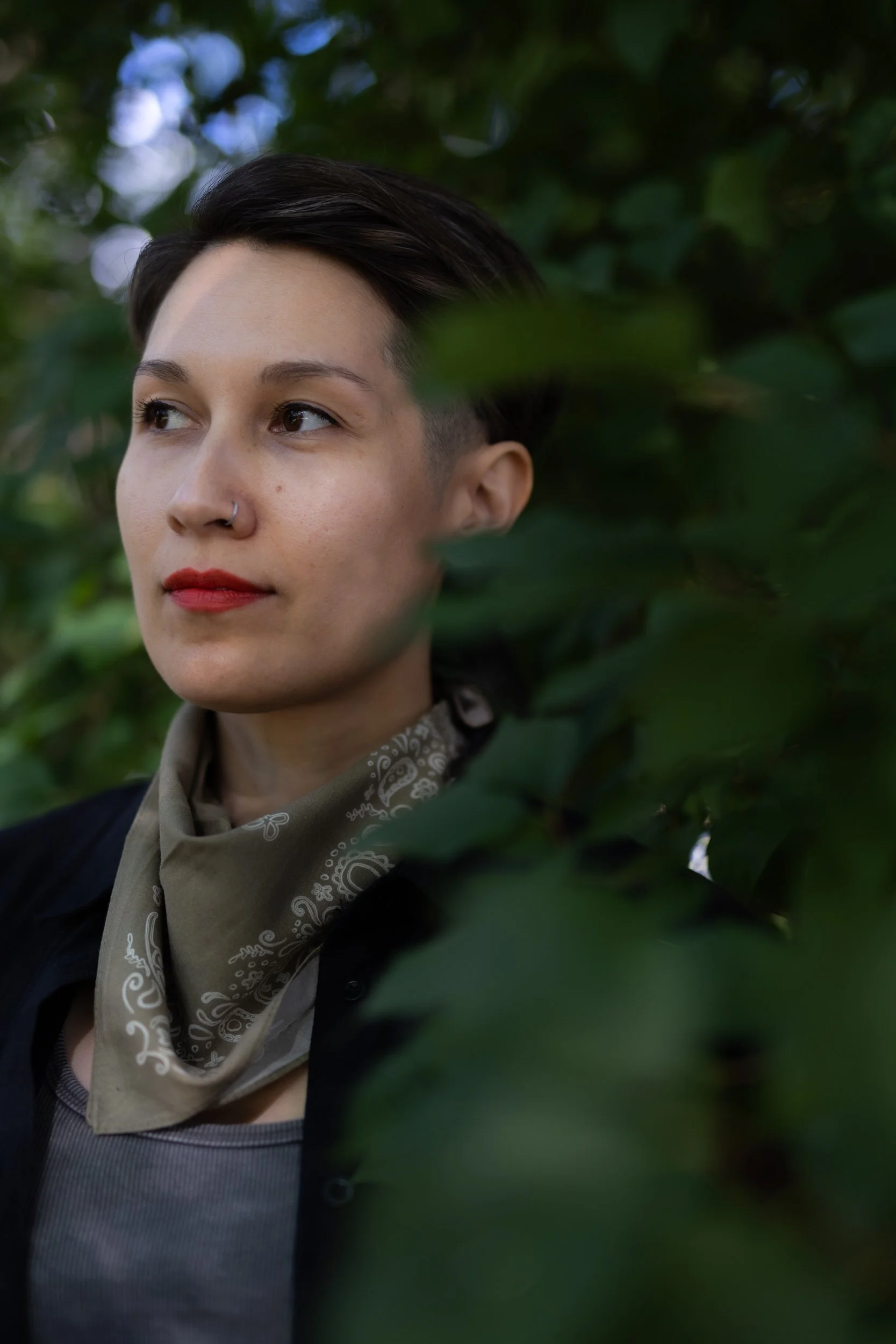 Close-up of a person with short dark hair, red lipstick, wearing a nose ring, and a beige bandana around their neck, surrounded by green leaves.