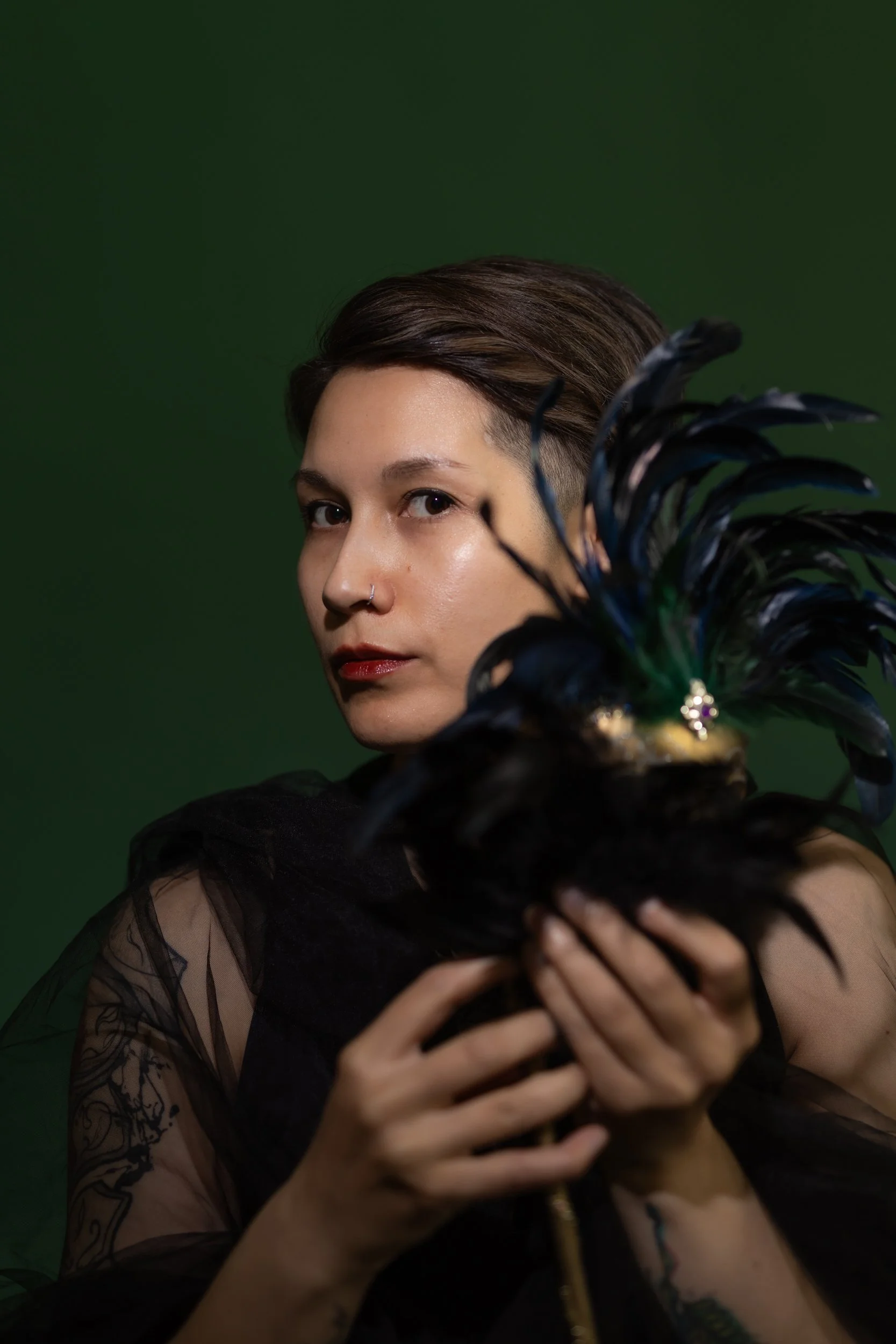 A person with short brown hair and a nose piercing holds a large peacock feather fan in front of their face, wearing a black outfit with sheer fabric and lace details, against a dark green background.