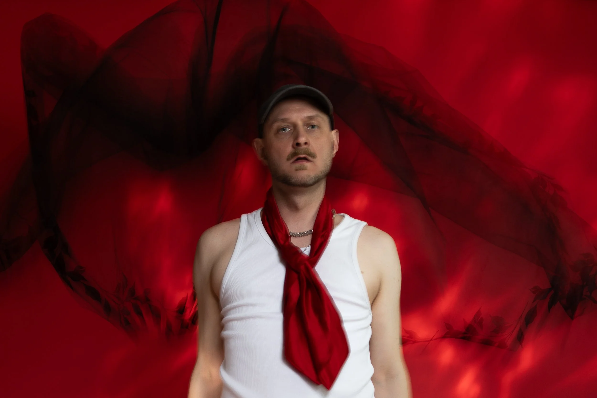 A person stands in front of a red background. Light falls on the backdrop, making it look as though it is on fire. Behind the person, a black veil is falling through the air, its movement captured as motion blur. They are wearing a white shirt.