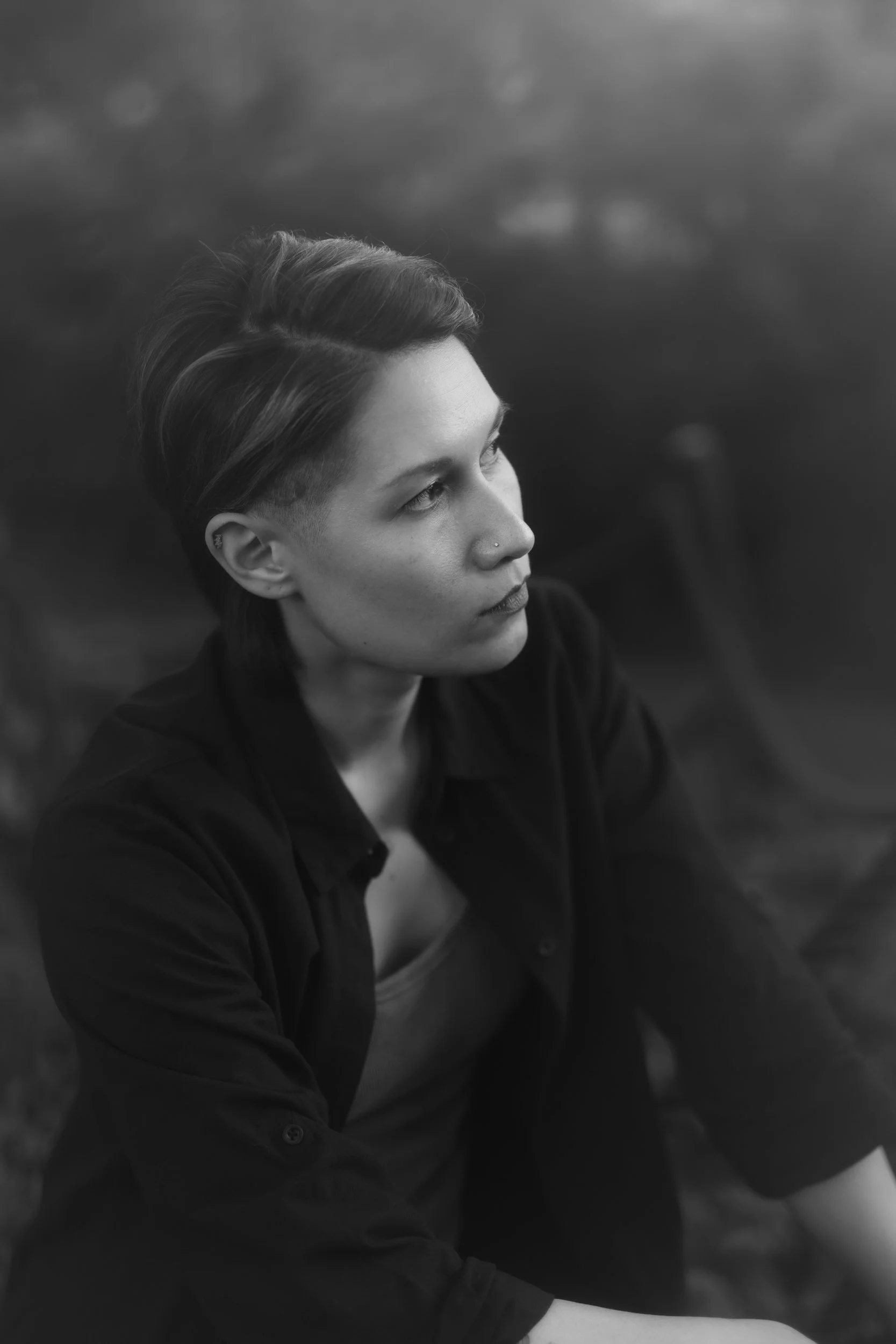 A black and white photo of a person with short hair, pierced nose, and earrings, looking to the right with a thoughtful expression. They are wearing a button-up shirt and are outdoors with a blurred natural background.