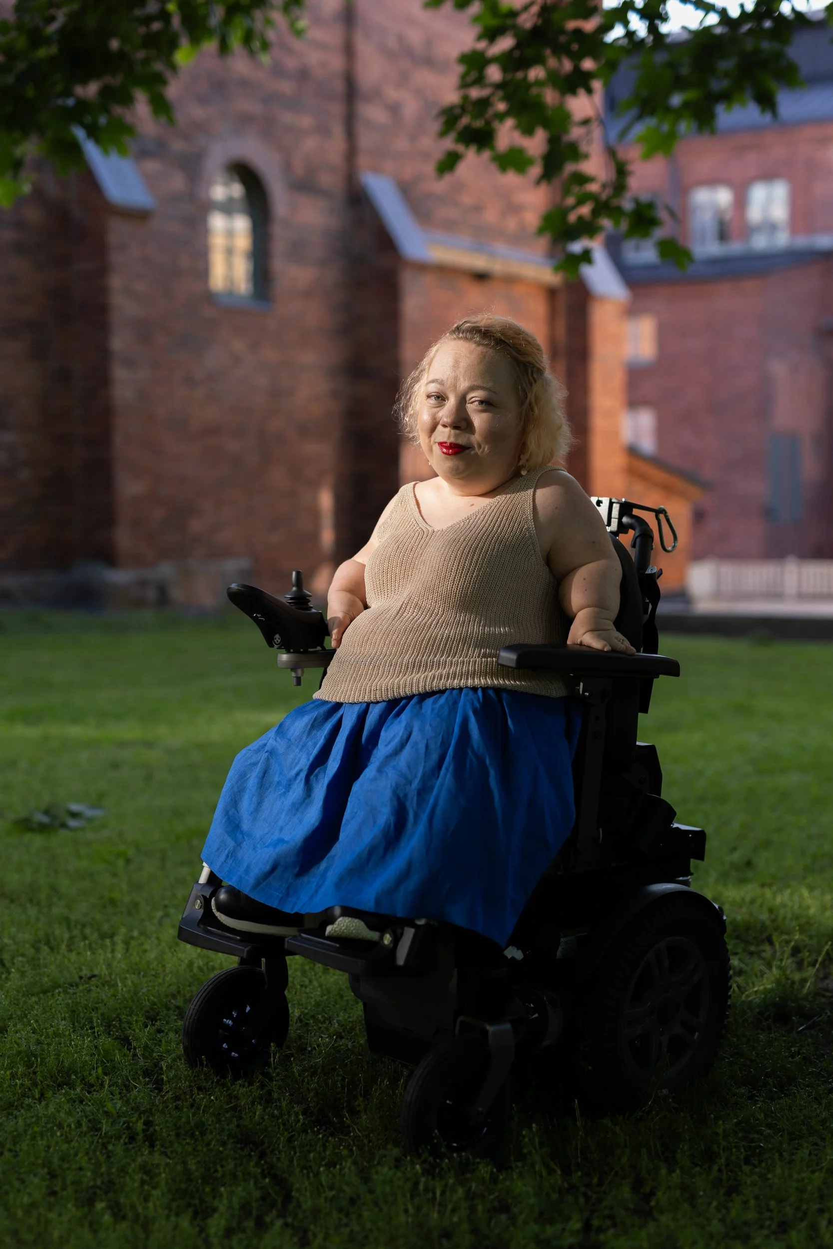 A short statured woman is sitting on a wheelchair. She is smiling and looking happy. It is summer and she is wearing a blue skirt and beige knitted top.