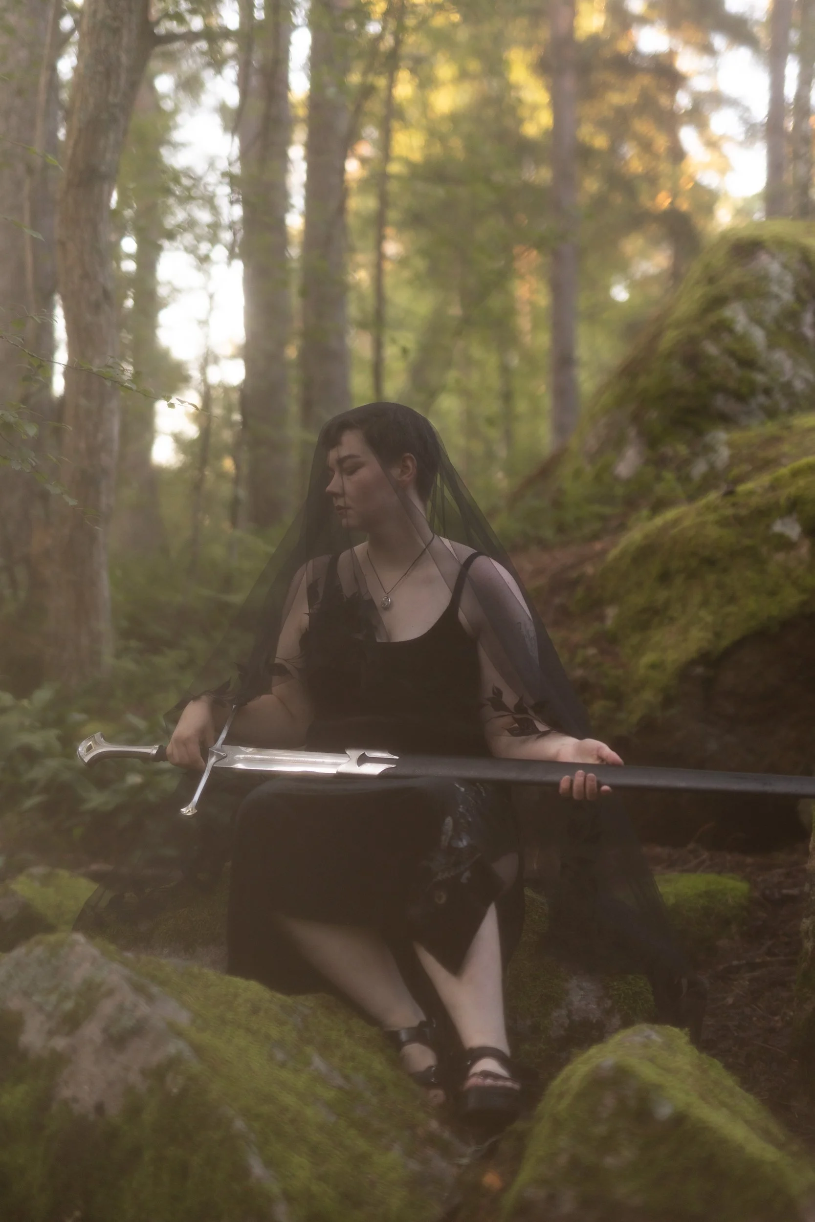 A person with black hair wearing a black dress and veil, sitting on a rockin a mossy forest setting with trees and soft sunlight in the background. They are holding a sword on their lap.