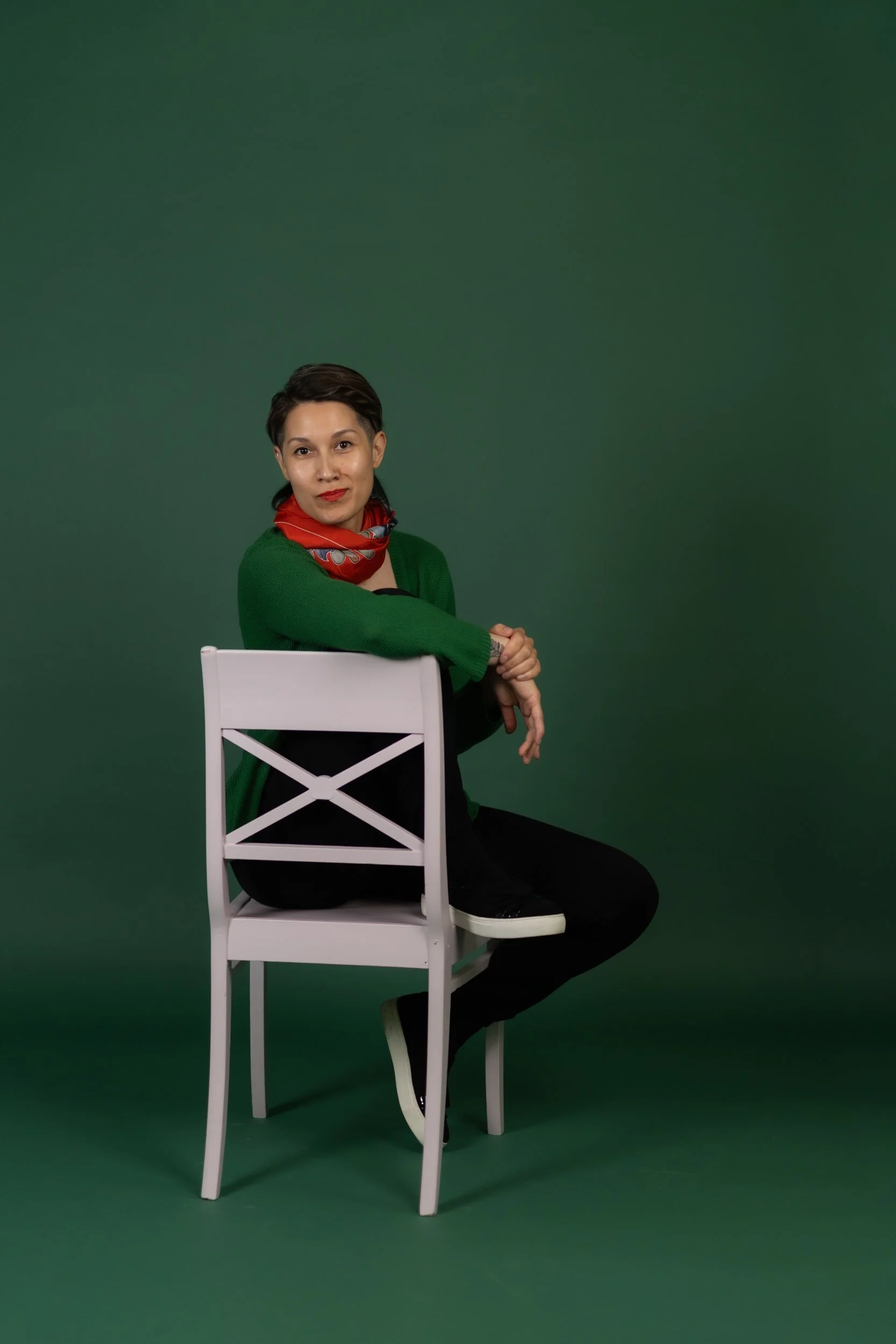 A person with short dark hair sitting on a white chair against a green background, wearing a red scarf, green cardigan, black shirt, and black pants.
