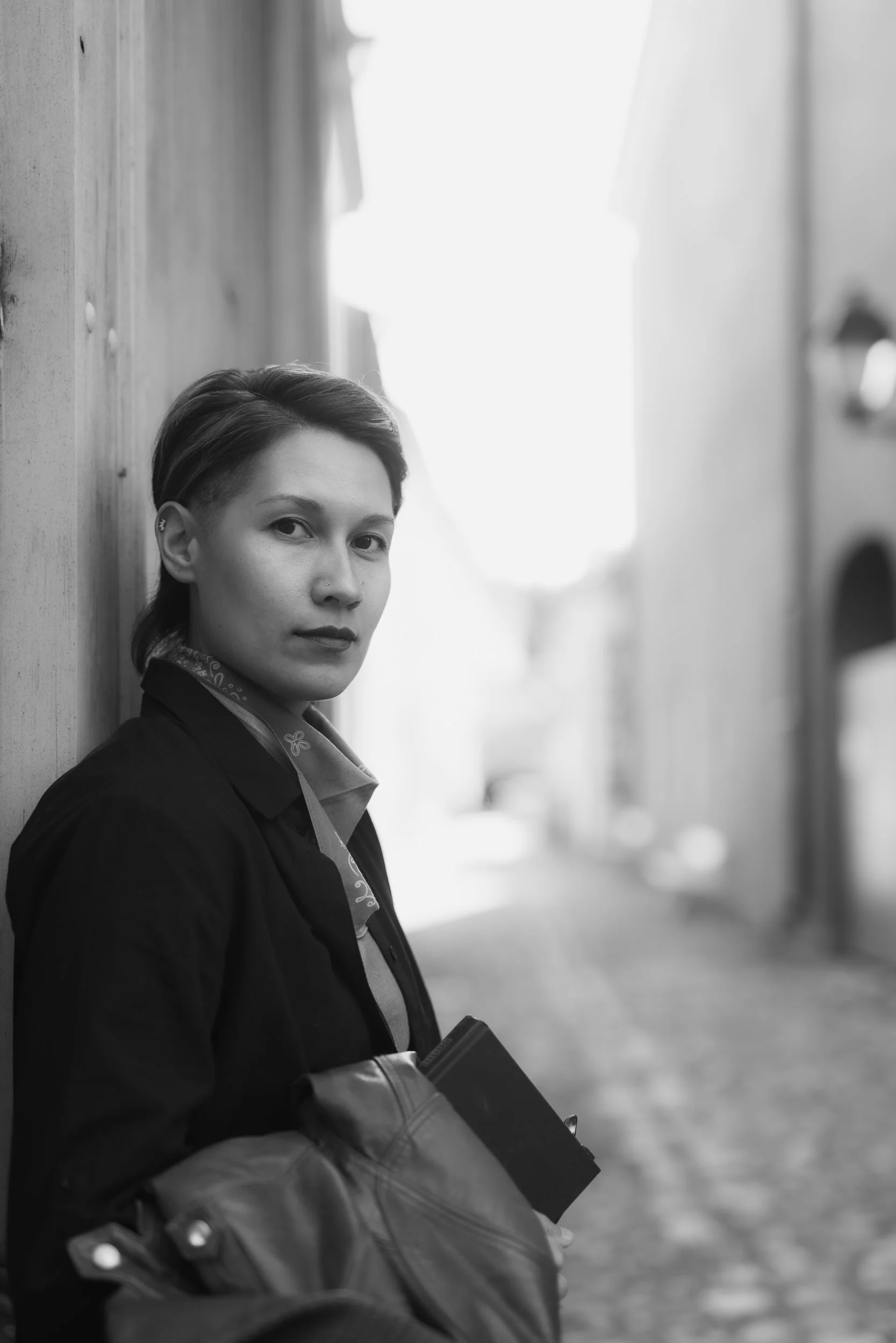 A person with short dark hair, wearing a black jacket and gloves, leaning against a wall outdoors, holding a notebook or folder, with a blurred background.