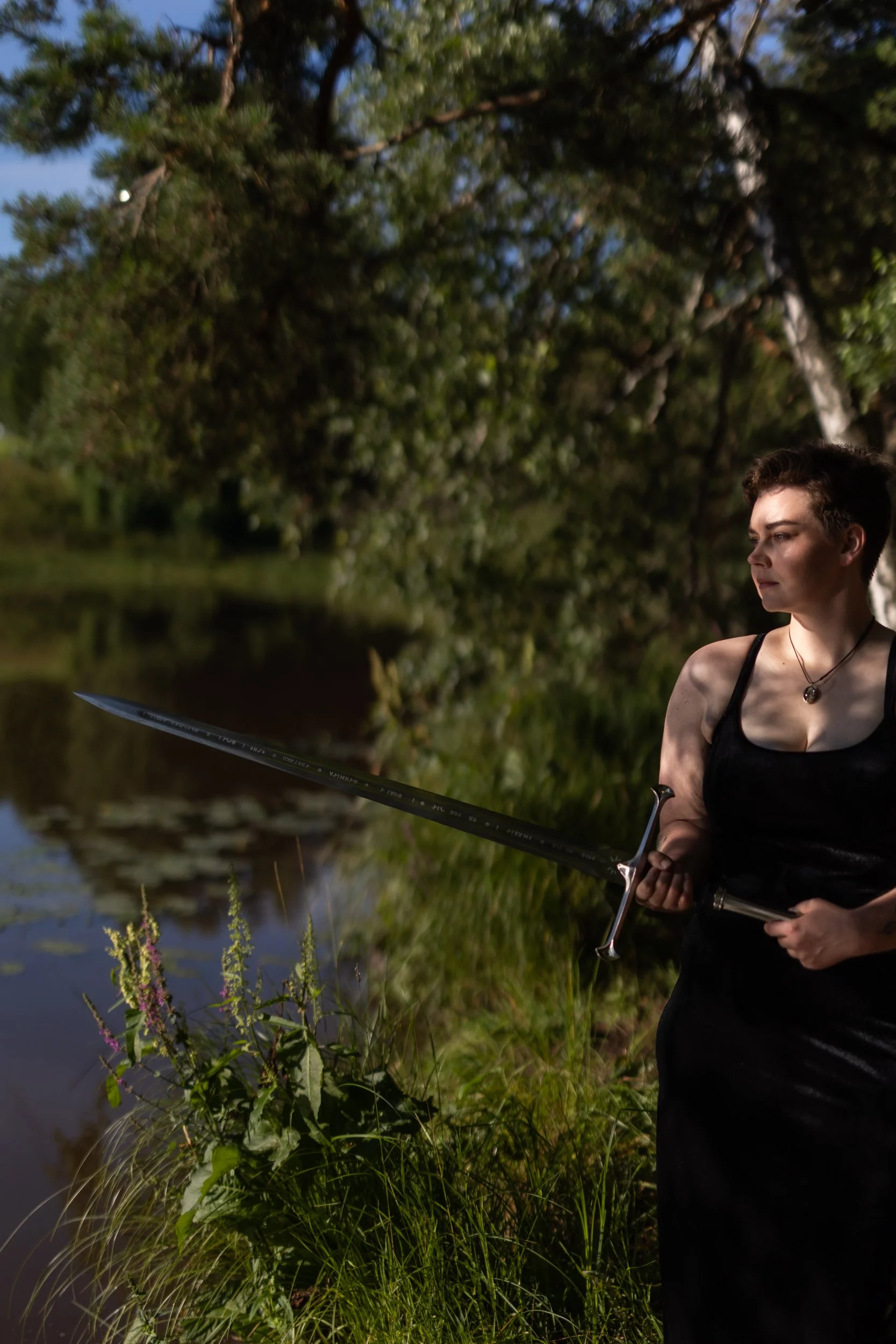 A person with short brown hair dressed in a black shirt with spaghetti straps, holding a bow and arrow, standing near a body of water surrounded by greenery and trees, during daytime.