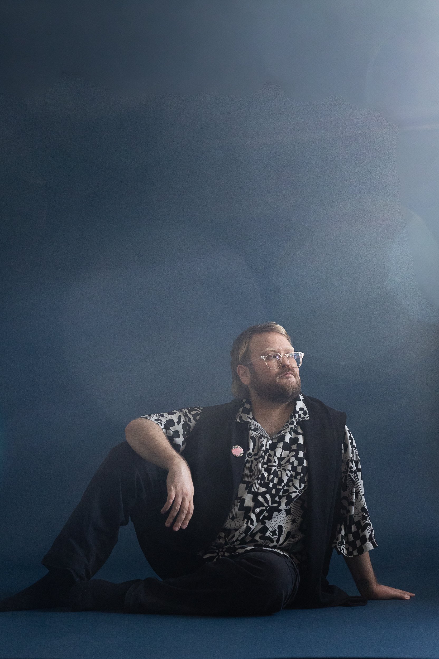Artist Taneli Kemppi is sitting on a dark blue backdrop. Sun is shining and there a les flares that make the image hazy. Taneli is leaning on their left hand and looking past the camera.
